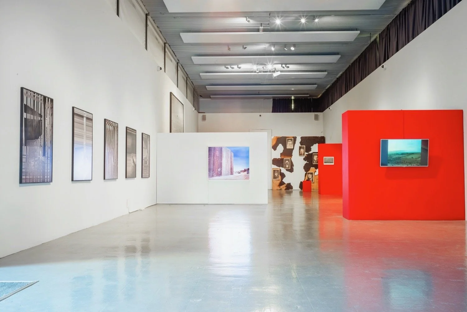 An art gallery with framed photographs on white walls, a large photograph on a white partition wall, and various textured wall displays. There is a red display with a mounted screen showing a landscape. The gallery has a high ceiling with track light