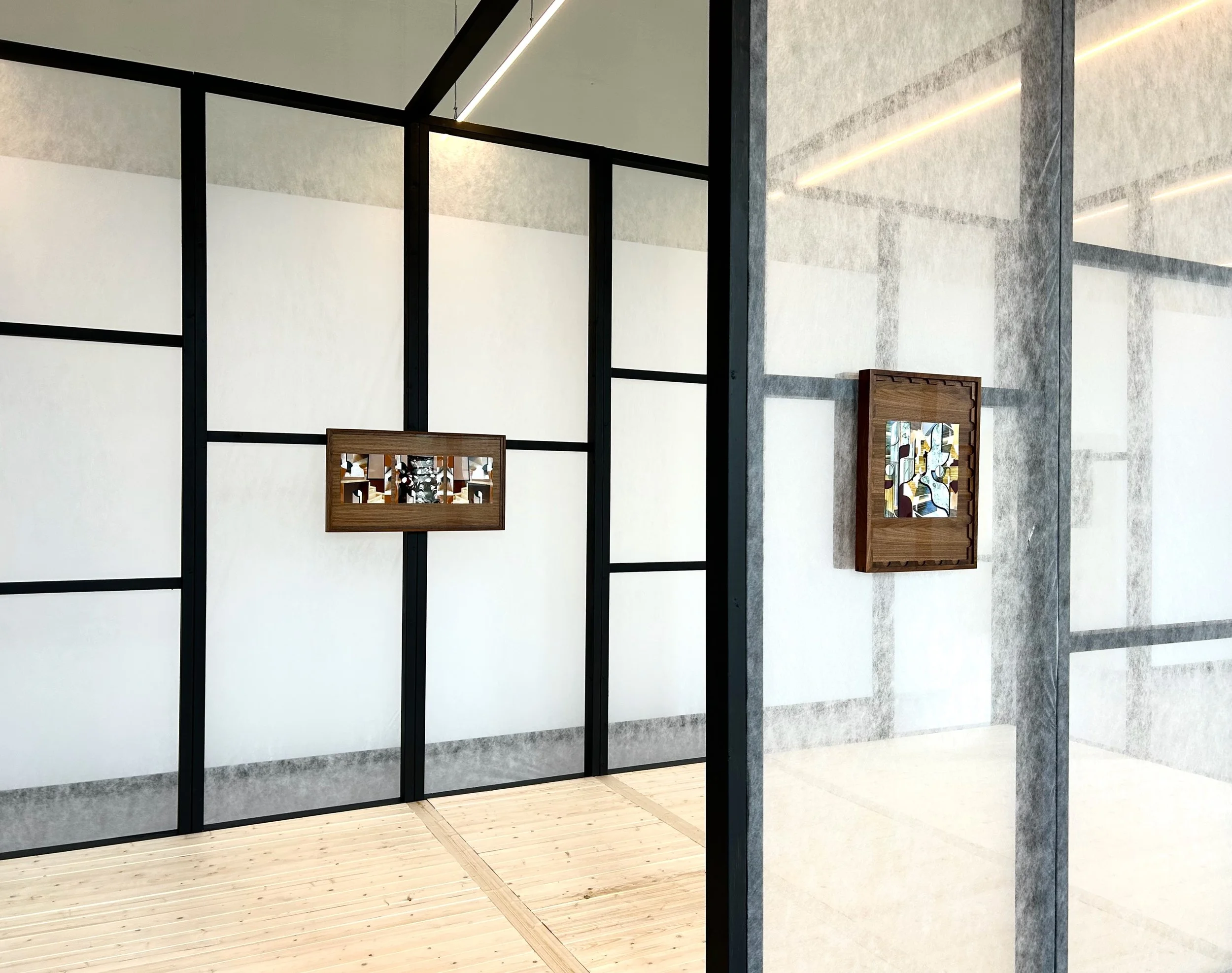 Interior view of a modern art gallery with white walls, black framing, wooden flooring, and artworks displayed in wooden frames.