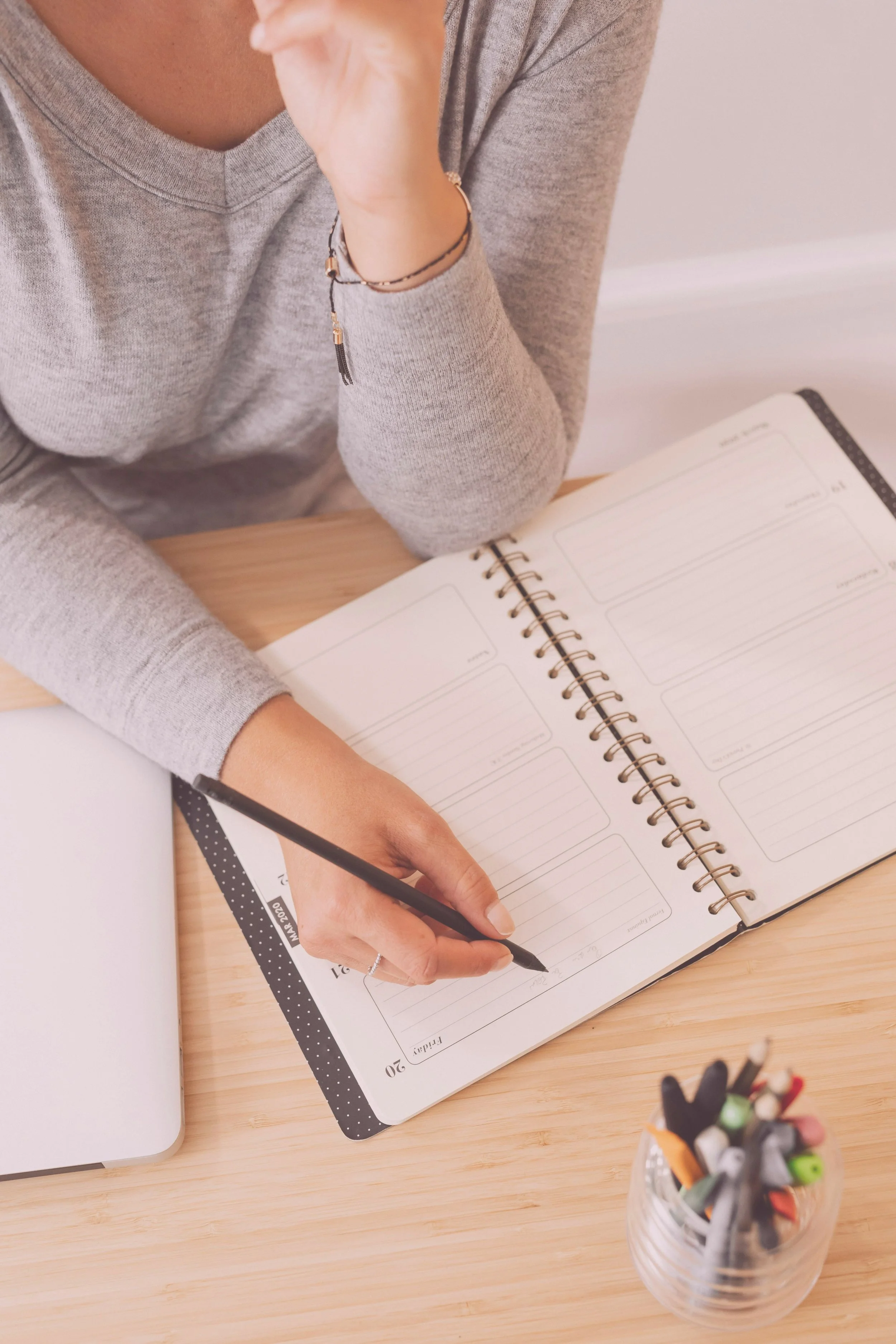 Person writing in a planner at a desk with a container of colored pens nearby.