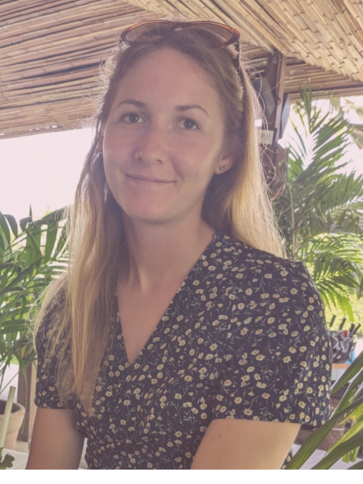 A woman with long blonde hair wearing a black floral dress, sitting in a tropical outdoor setting with green plants and a bamboo roof.