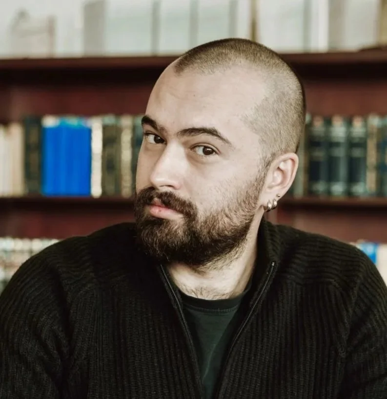 A man with a beard, mustache, and shaved head, wearing earrings, looking at the camera in a library.