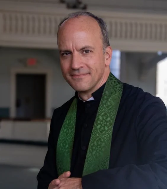 A middle-aged man dressed in a black clergy robe with a green stole, standing indoors with an elegant background.