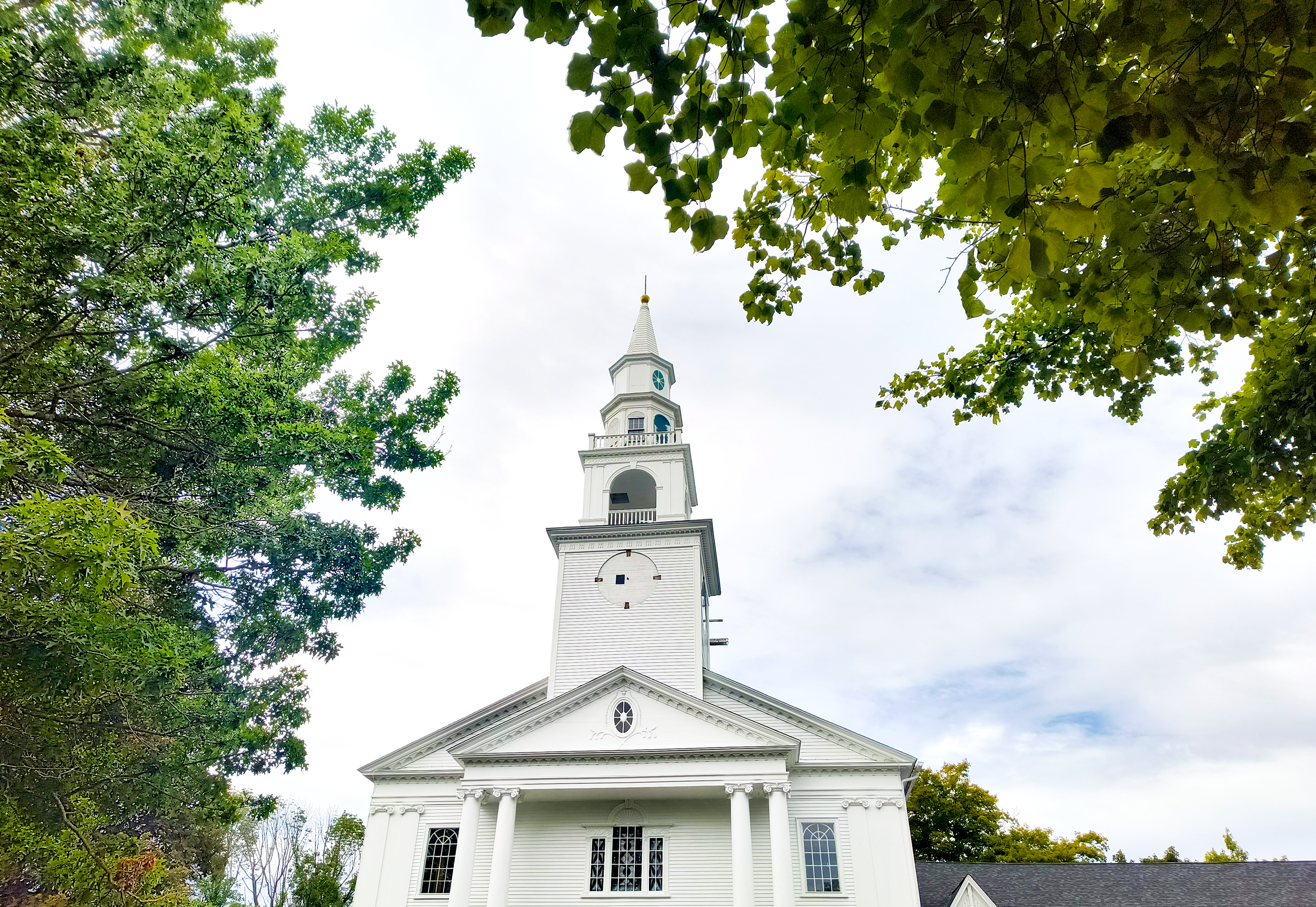 White church with steeple surrounded by green leaves and trees, cloudy sky in the background.