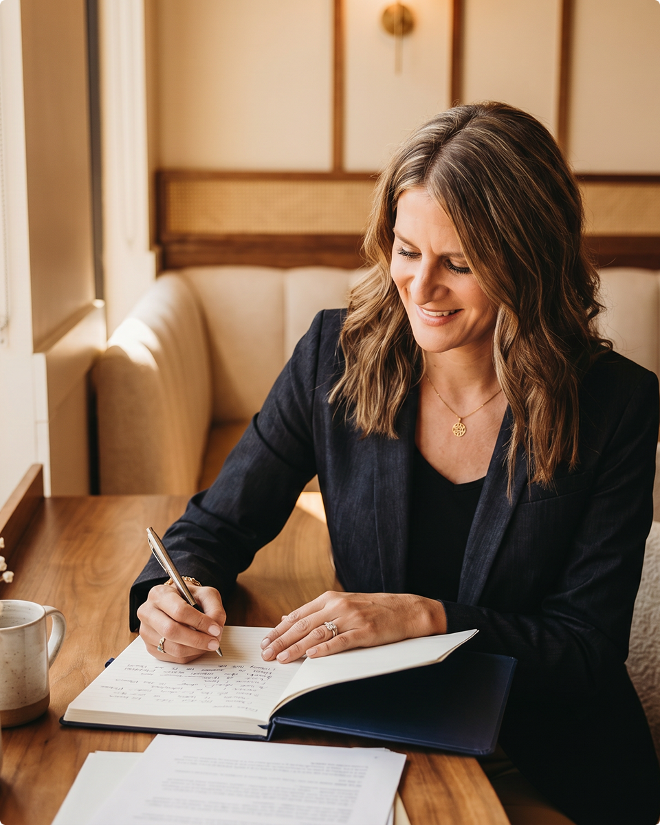 A woman with wavy brown hair writing in a notebook at a wooden table, smiling and wearing a black blazer and gold necklace in a cozy café setting.