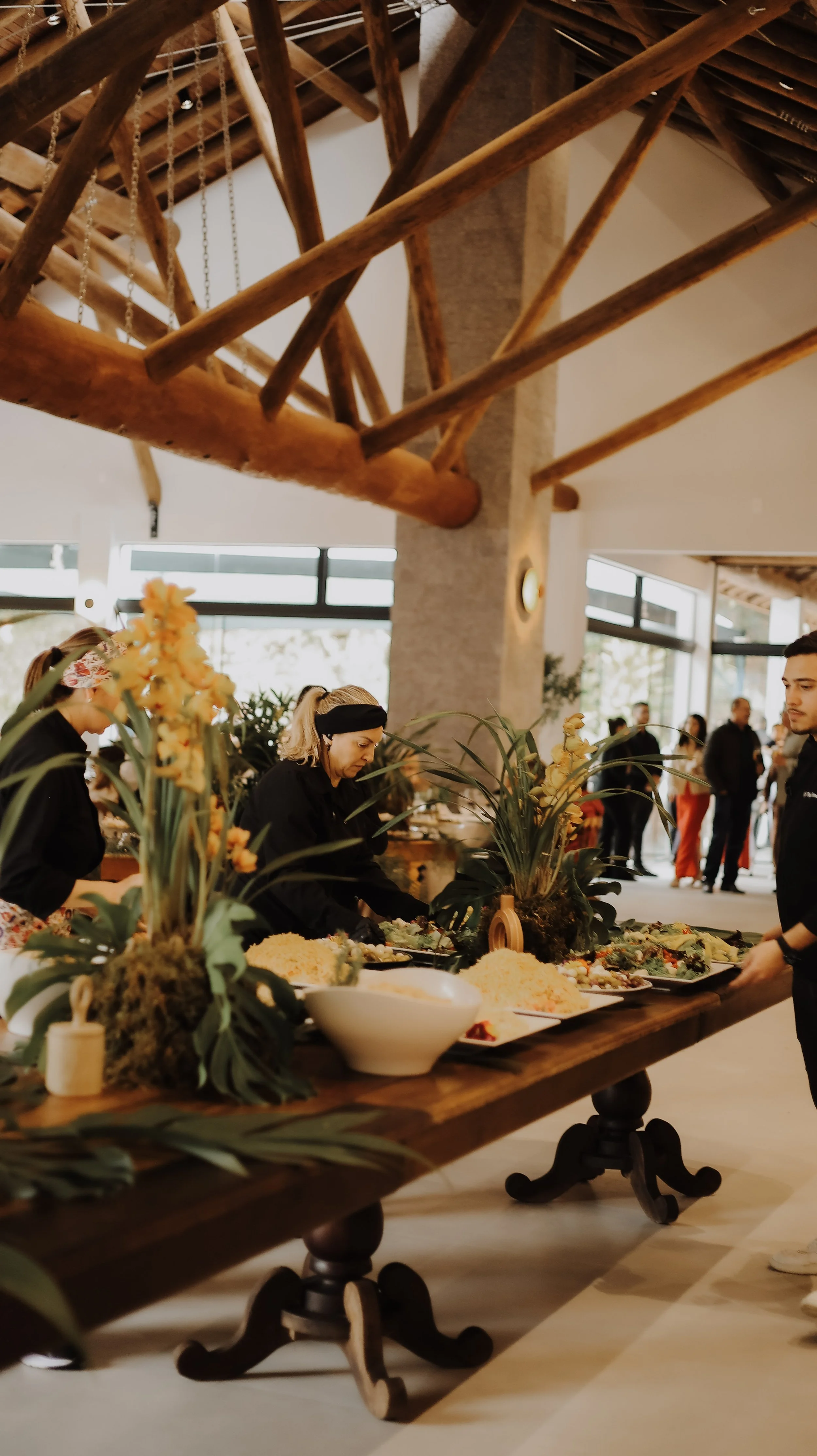 Buffet de comida com várias pessoas ao fundo, decorado com plantas e arranjos florais em uma sala de edifício com janelas grandes e estrutura de madeira visível no teto.