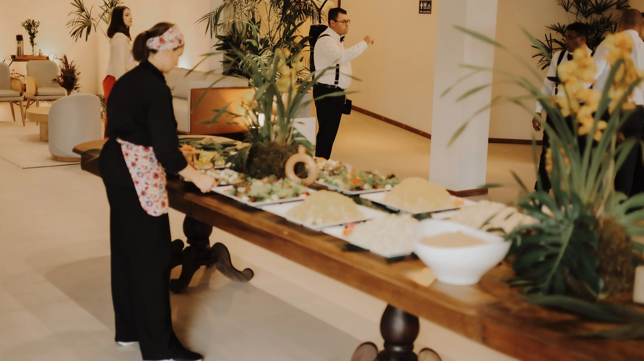 Funcionários em uniforme de um restaurante de luxo preparando uma mesa de buffet com pratos decorados, enquanto outros funcionários ajustam a decoração na sala.