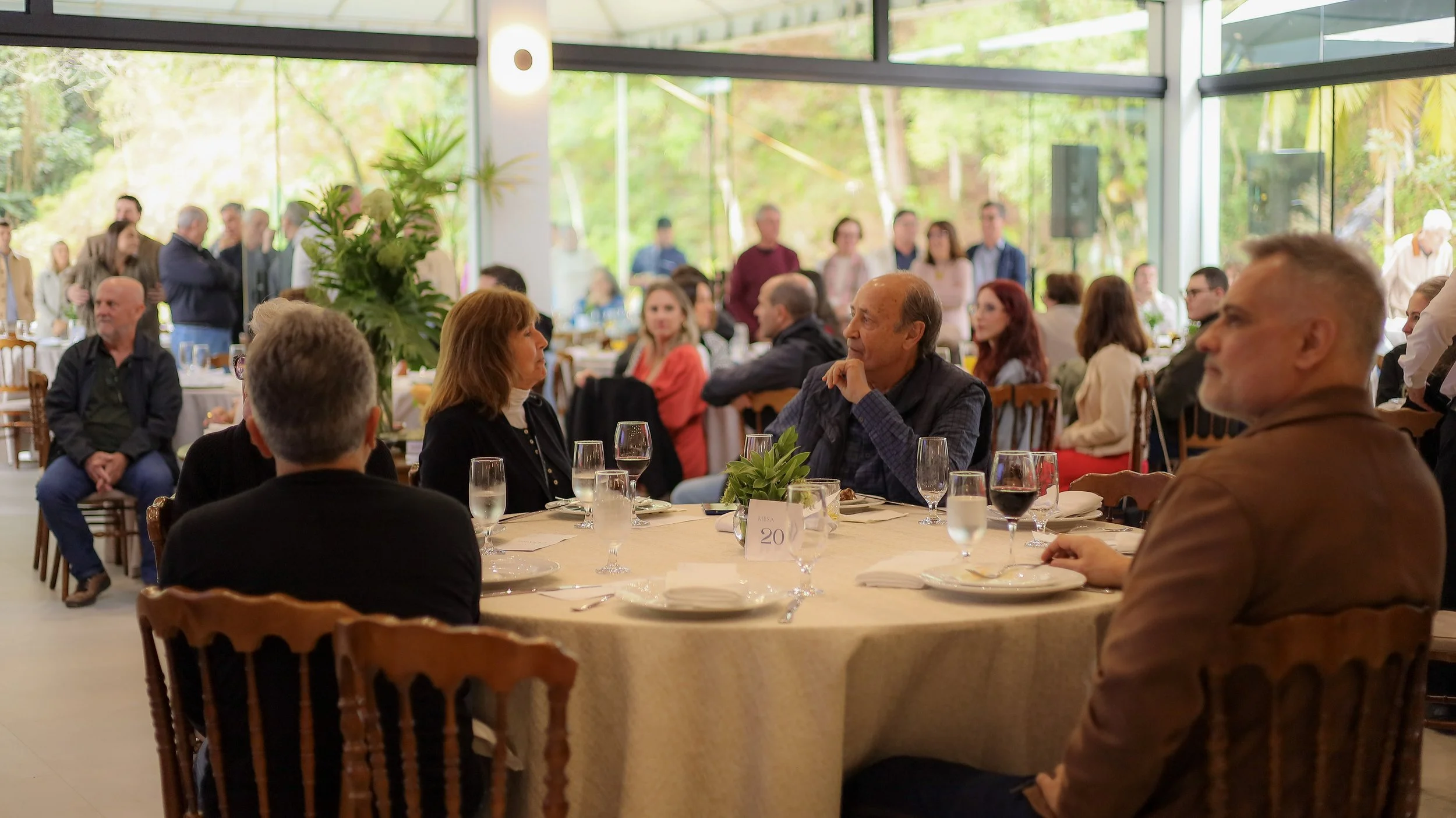 Pessoas sentadas ao redor de uma mesa em um evento social, com janelas amplas e vista para um ambiente ao ar livre com árvores e plantas.