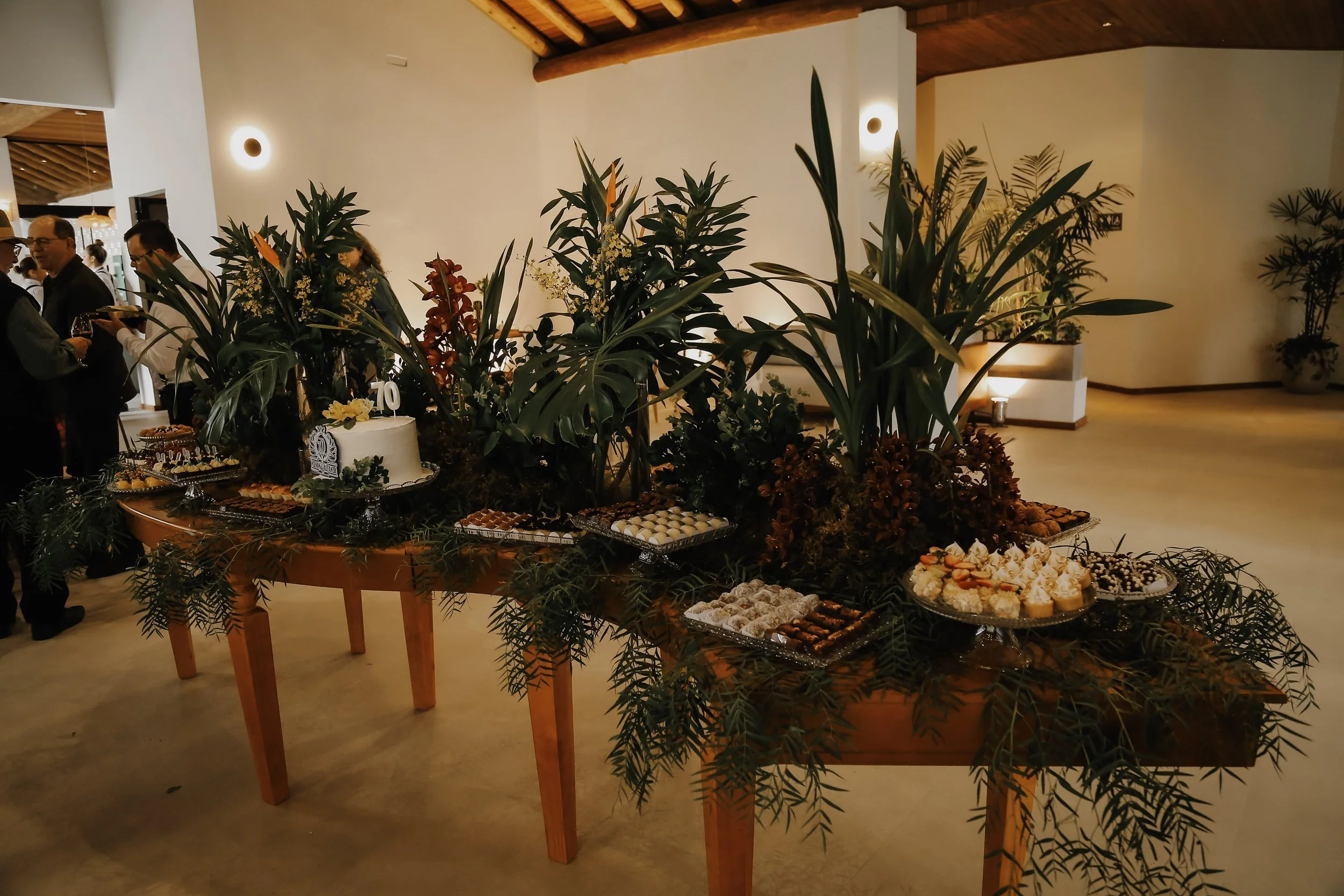 Mesa de bolos e doces decorada com plantas verdes e flores em festa ou celebração.