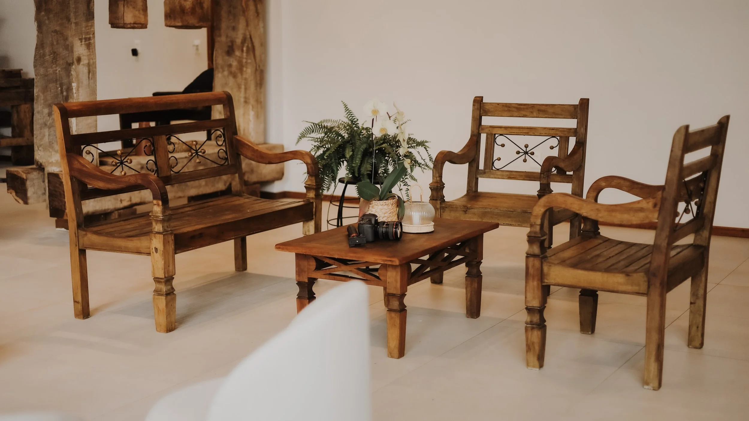 Sala com móveis de madeira, incluindo um sofá de dois lugares e duas cadeiras, uma mesa de centro com uma câmera e acessórios, uma planta em vaso no centro da sala, e uma parede branca ao fundo.