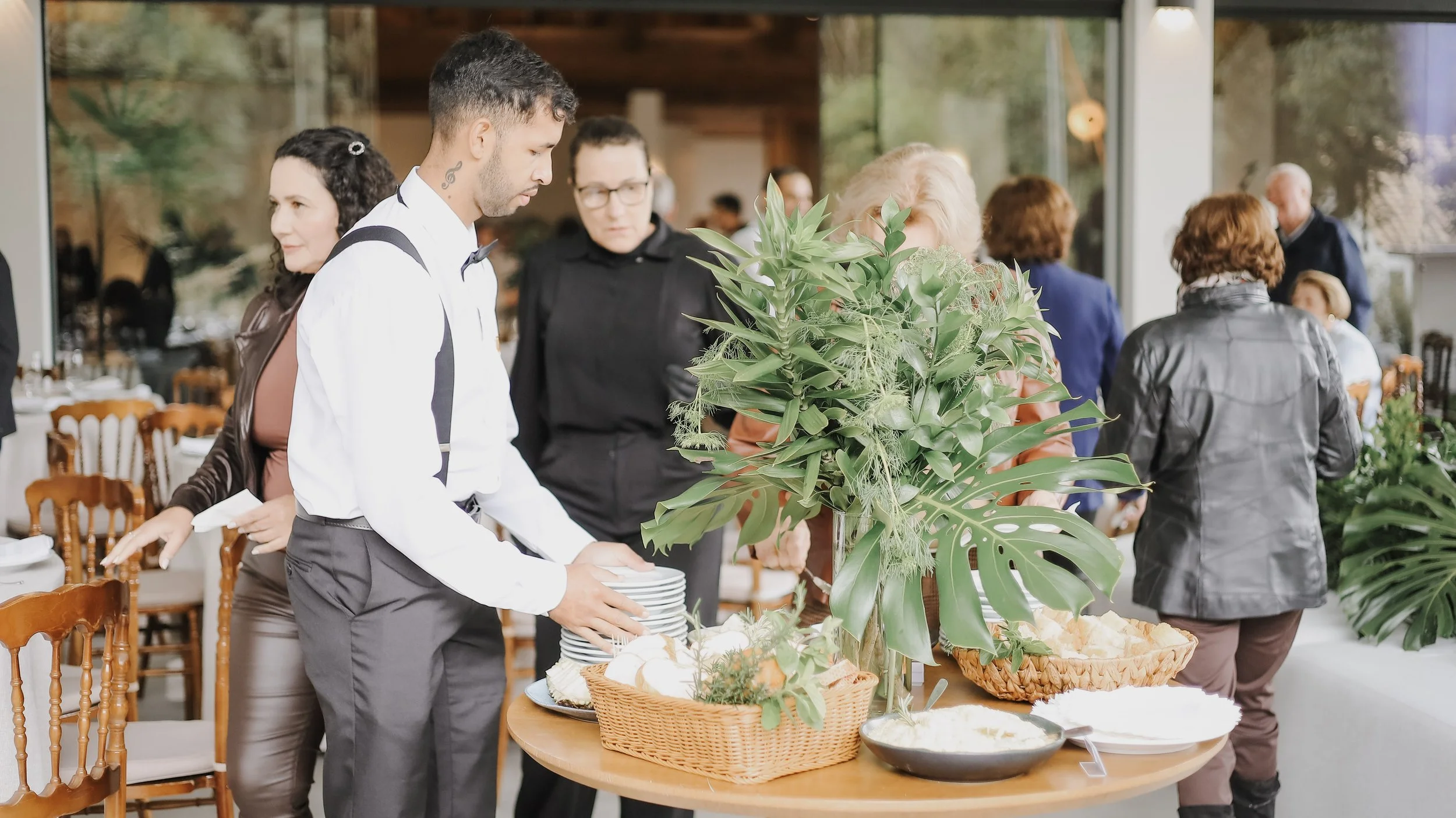 Pessoas se servindo de comida em uma mesa com plantas verdes decorativas em um evento ou buffet.