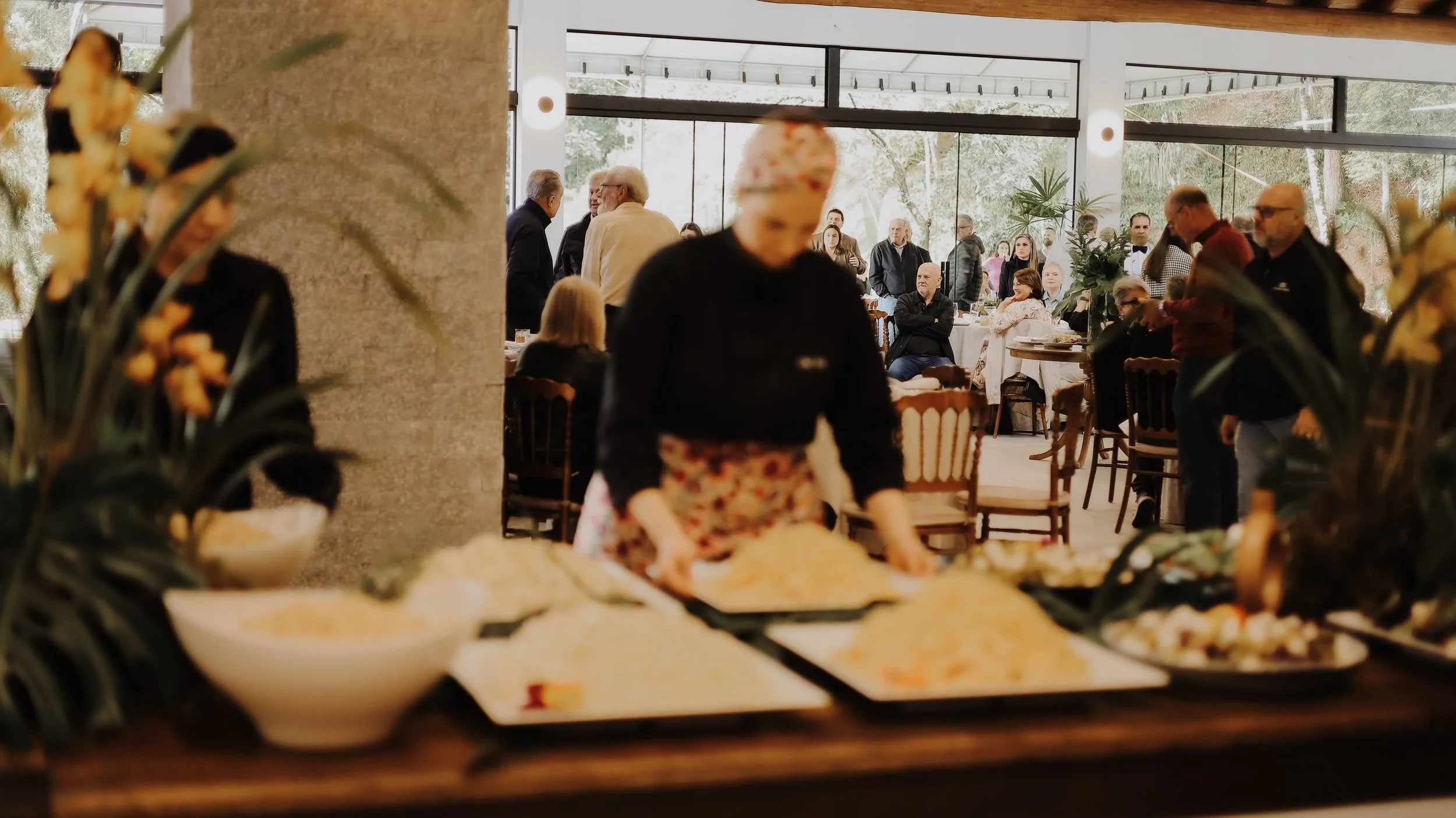 Vista de uma sala de banquete com pessoas conversando ao redor de mesas, com uma pessoa ao fundo preparando comida na mesa de bufê, decorada com flores e plantas.
