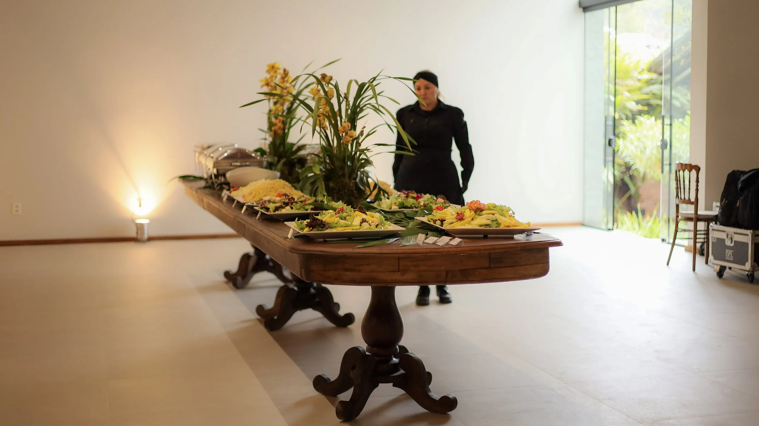 Mesa de buffet com saladas e pratos variados, decorada com plantas e flores, em um ambiente bem iluminado com acesso ao jardim. Uma atendente vestida de preto observa ao fundo.