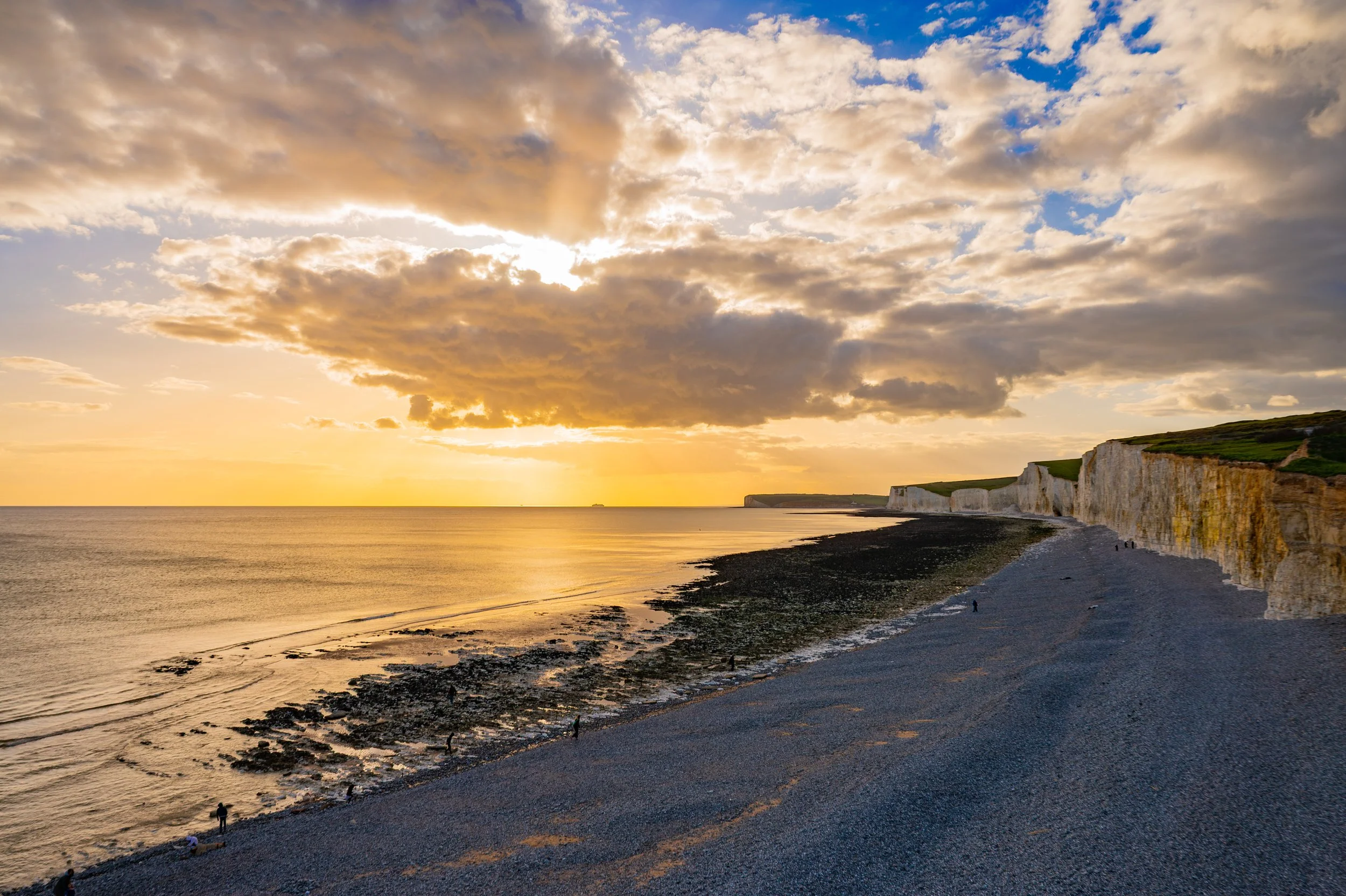 Scenic view of a coastline at sunset with white chalk cliffs towering over a pebble beach, calm water, and partly cloudy sky with golden sunlight.