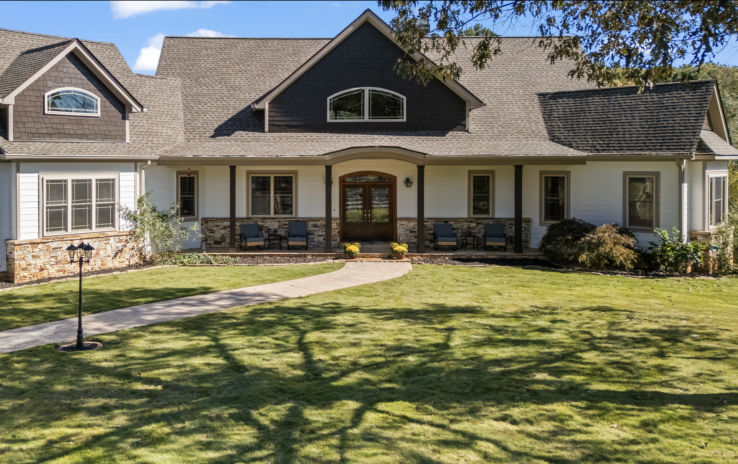 Front view of a large house with a brown shingled roof, white exterior walls, and a stone porch with four chairs. A curved concrete pathway leads through a well-manicured lawn to the front entrance, which is adorned with yellow flowers.