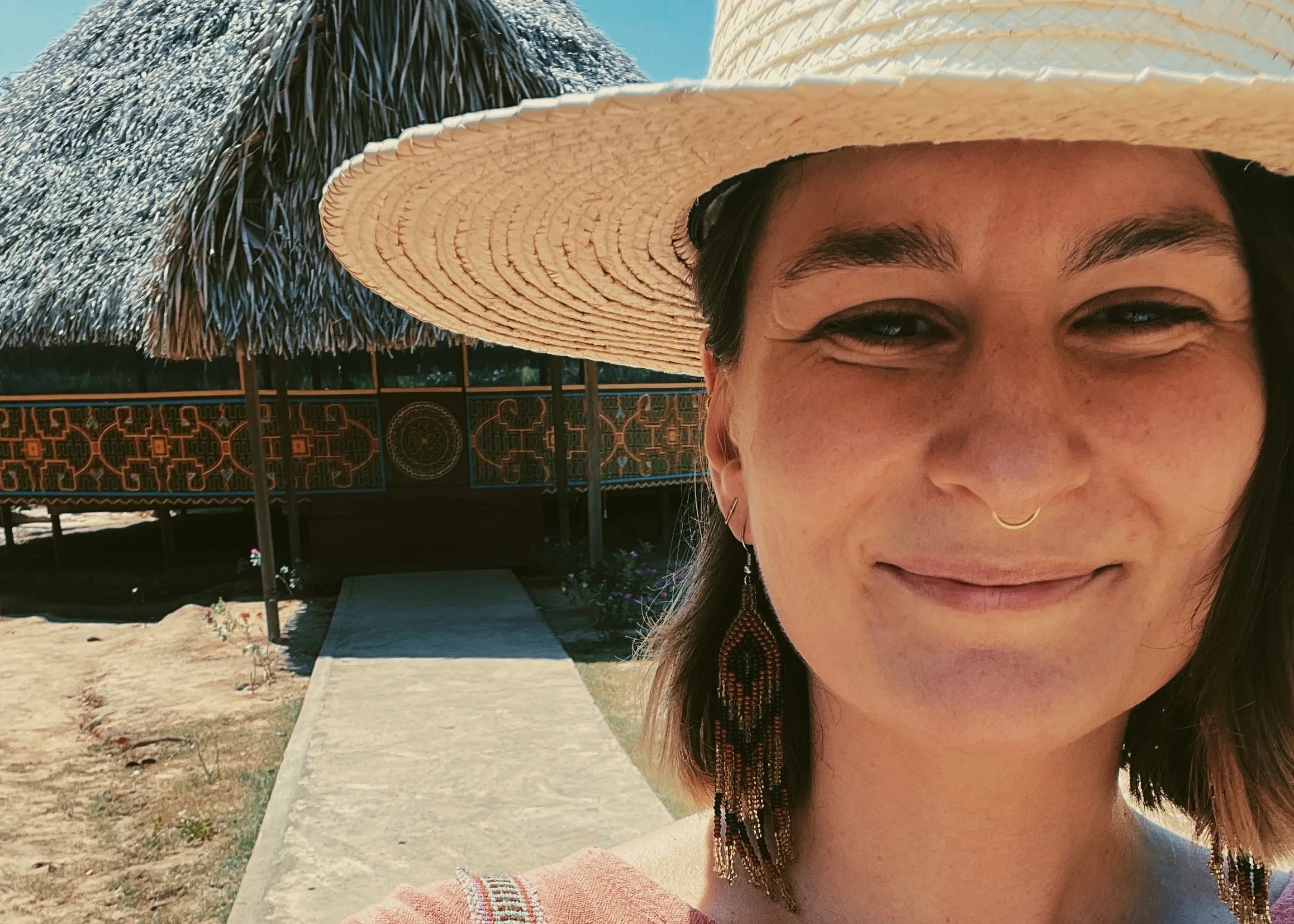 A woman with a hat, earrings, and a septum piercing smiling at the camera with a thatched-roof hut and bridge in the background.