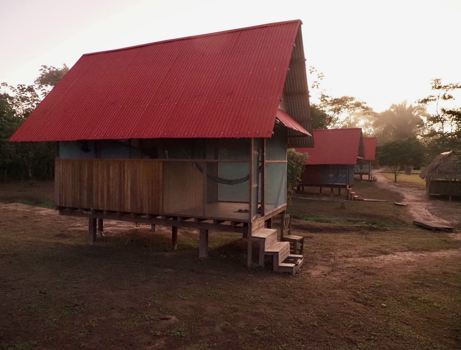A row of small, elevated houses with red metal roofs, surrounded by a dirt path and trees, with the sun setting in the background.