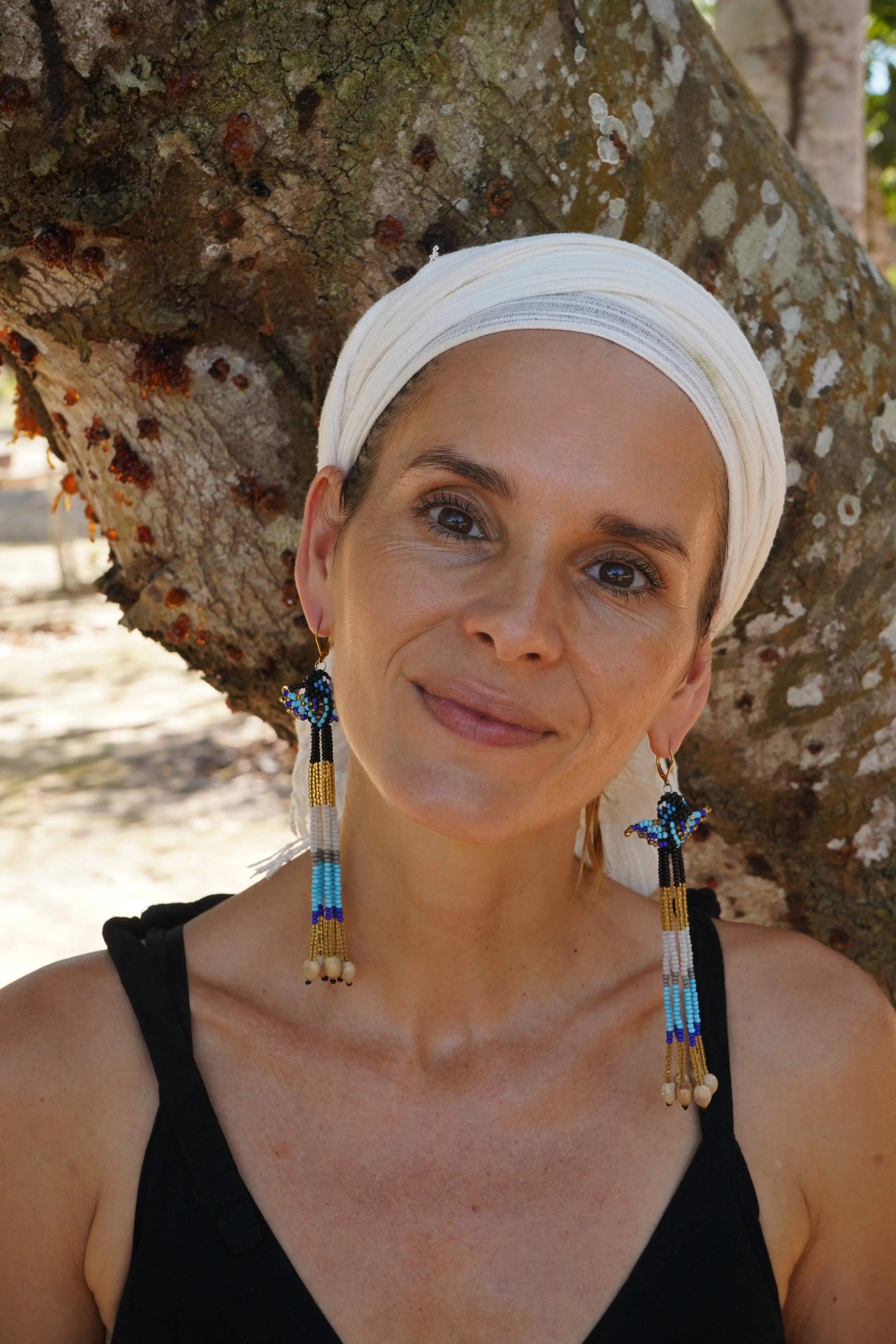 A woman with light skin, brown eyes, and blonde hair in a white headscarf, smiling while standing outdoors near a tree with textured bark and patches of lichen, wearing beaded earrings and a black sleeveless top.