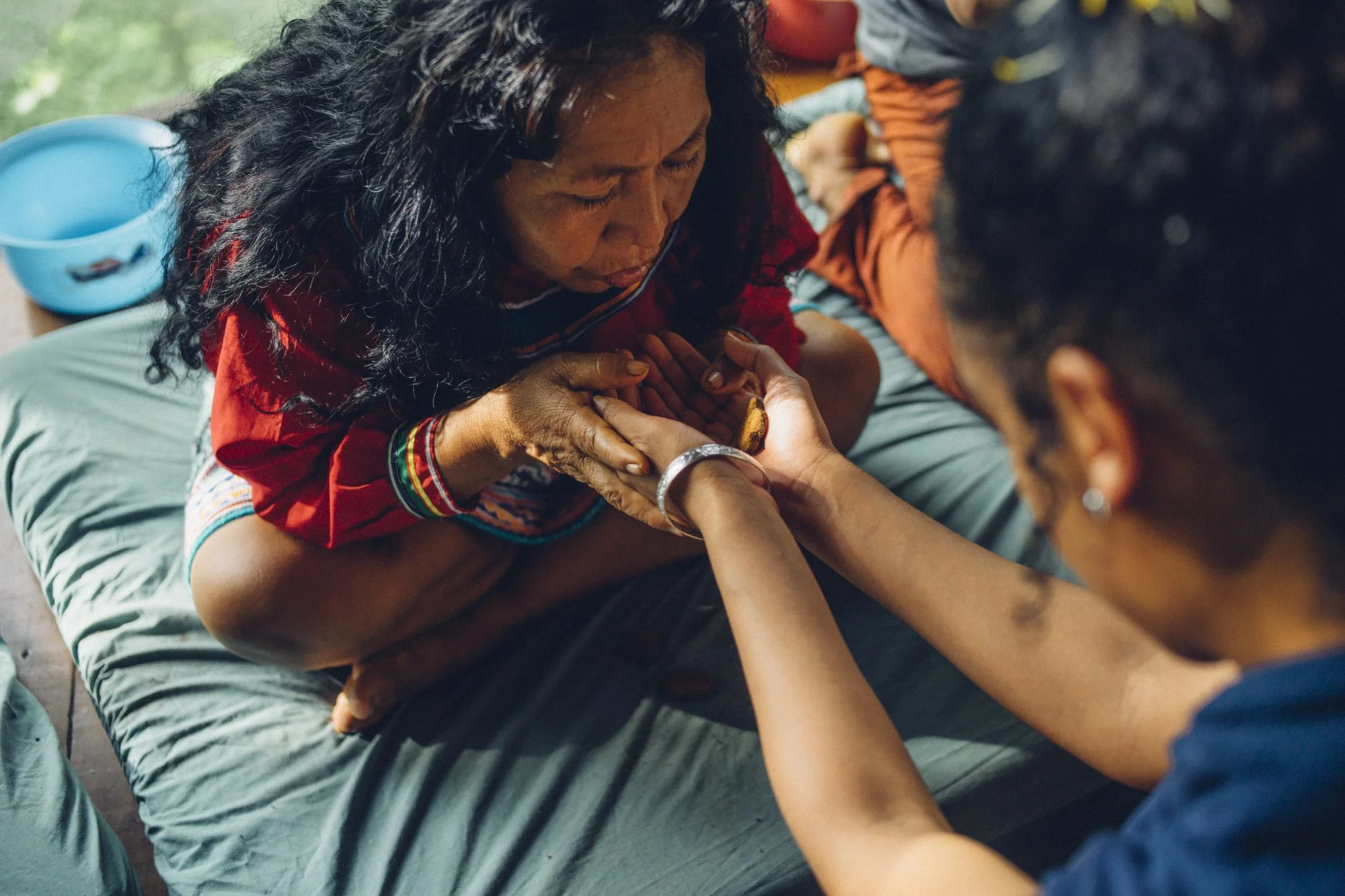 A woman with curly black hair and traditional clothing holds hands with a young girl, both sitting on a bed. The woman appears to be in a moment of prayer or reflection with her eyes closed. The young girl, who has dark hair tied back and is wearing a bracelet, is holding and touching the woman's hand. There is a blue plastic basin in the background.
