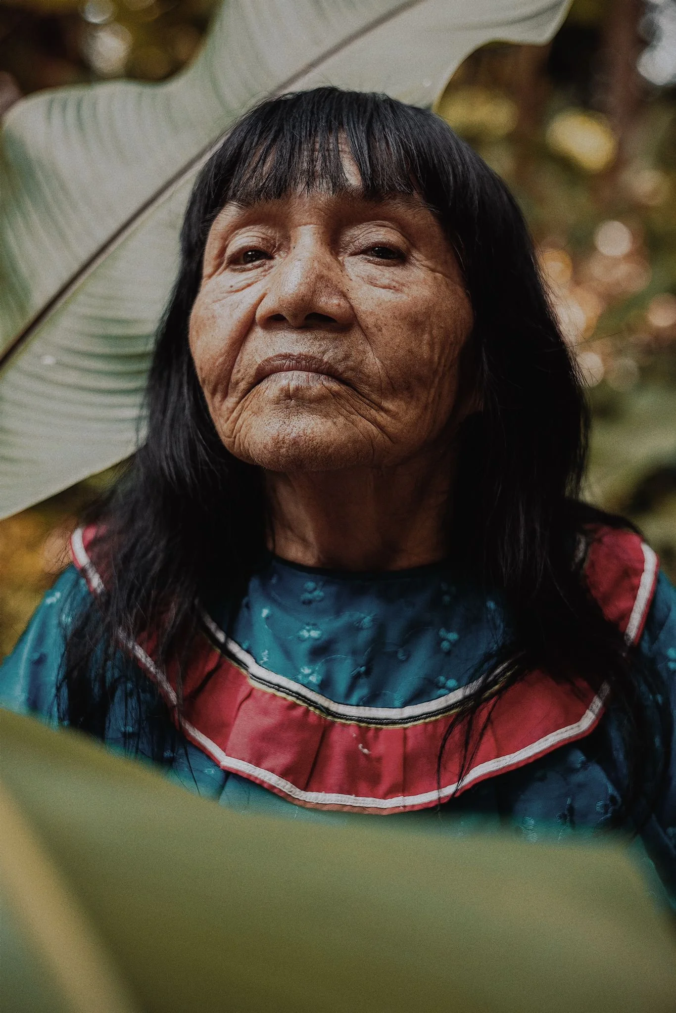 Close-up of an elderly woman with long black hair, wearing a colorful traditional garment, standing outdoors partially obscured by green leaves.