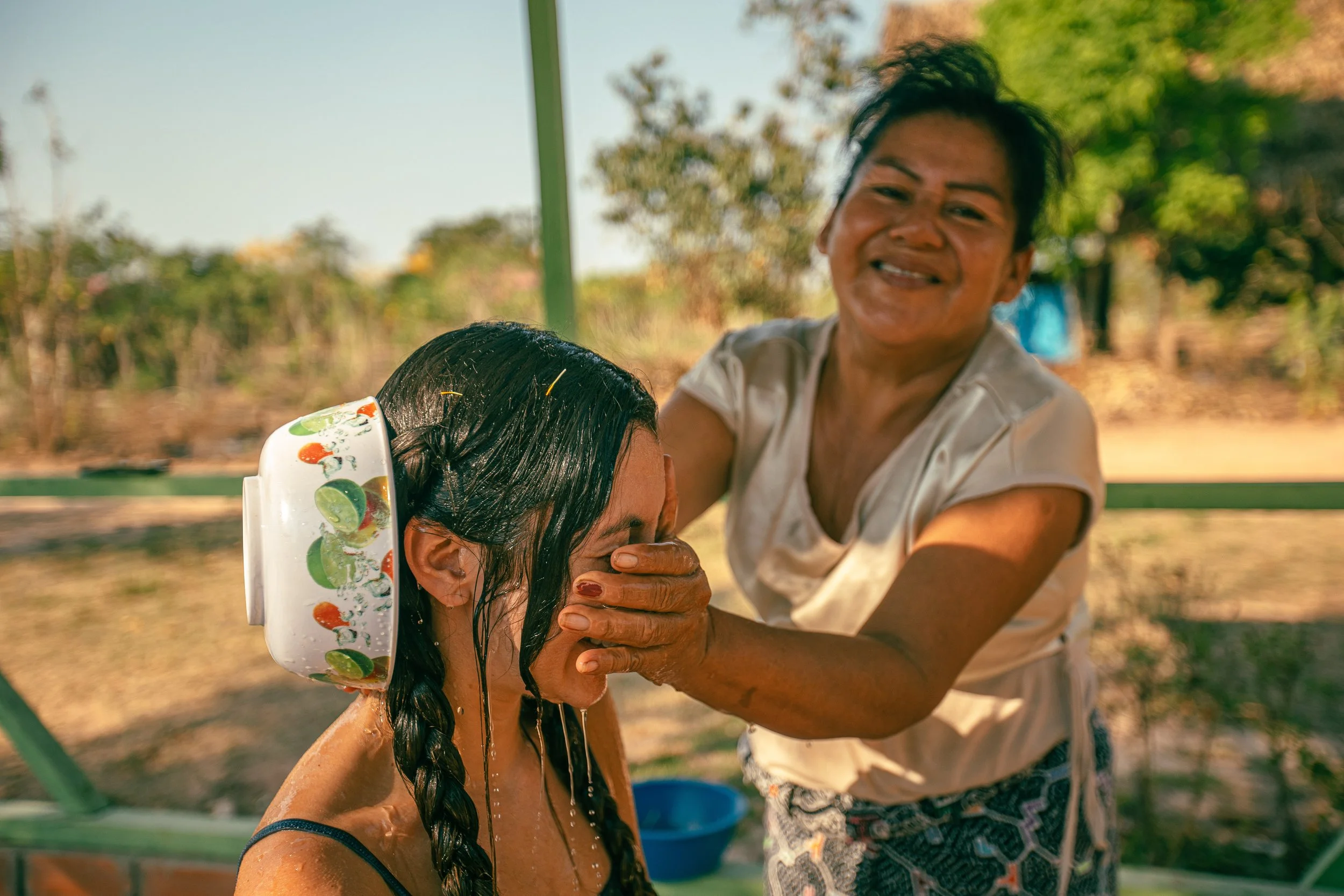 An older woman baptizing a young woman in an outdoor setting, with water splashing on her hair.