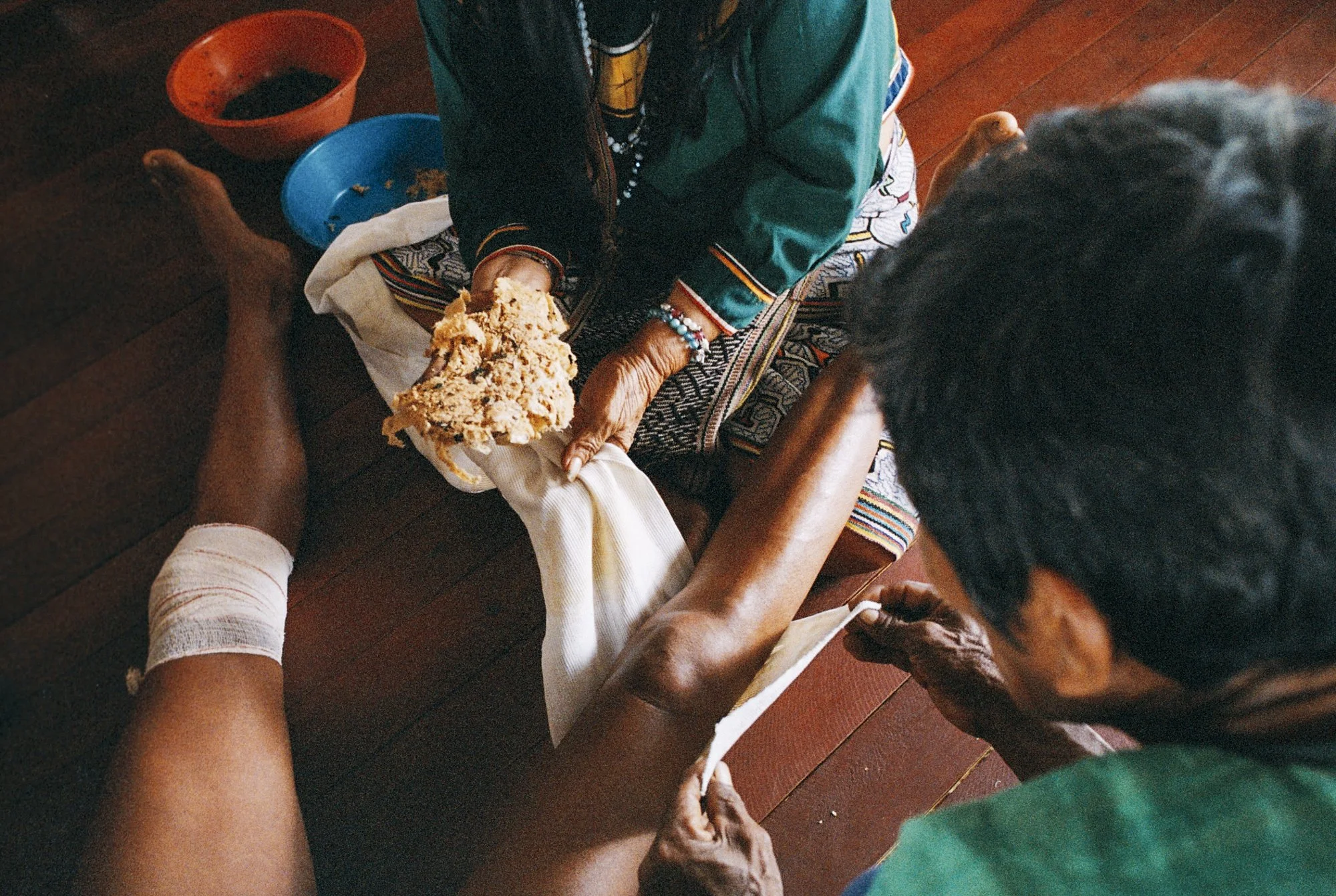 Older woman in colorful clothing petting a person's injured leg wrapped in a bandage, while seated on a wooden floor.