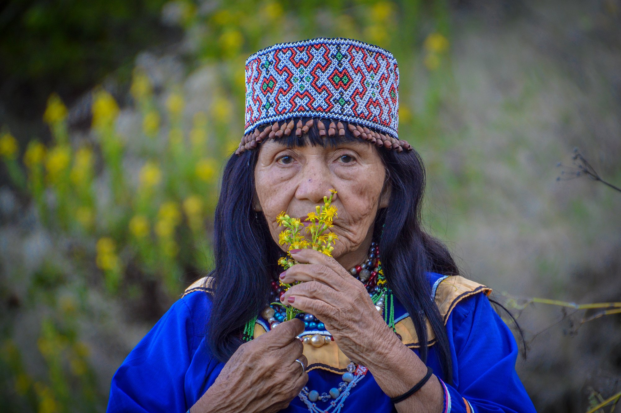 An elderly woman with long dark hair wearing traditional colorful clothing and a beaded hat, holding yellow flowers near her face outdoors.