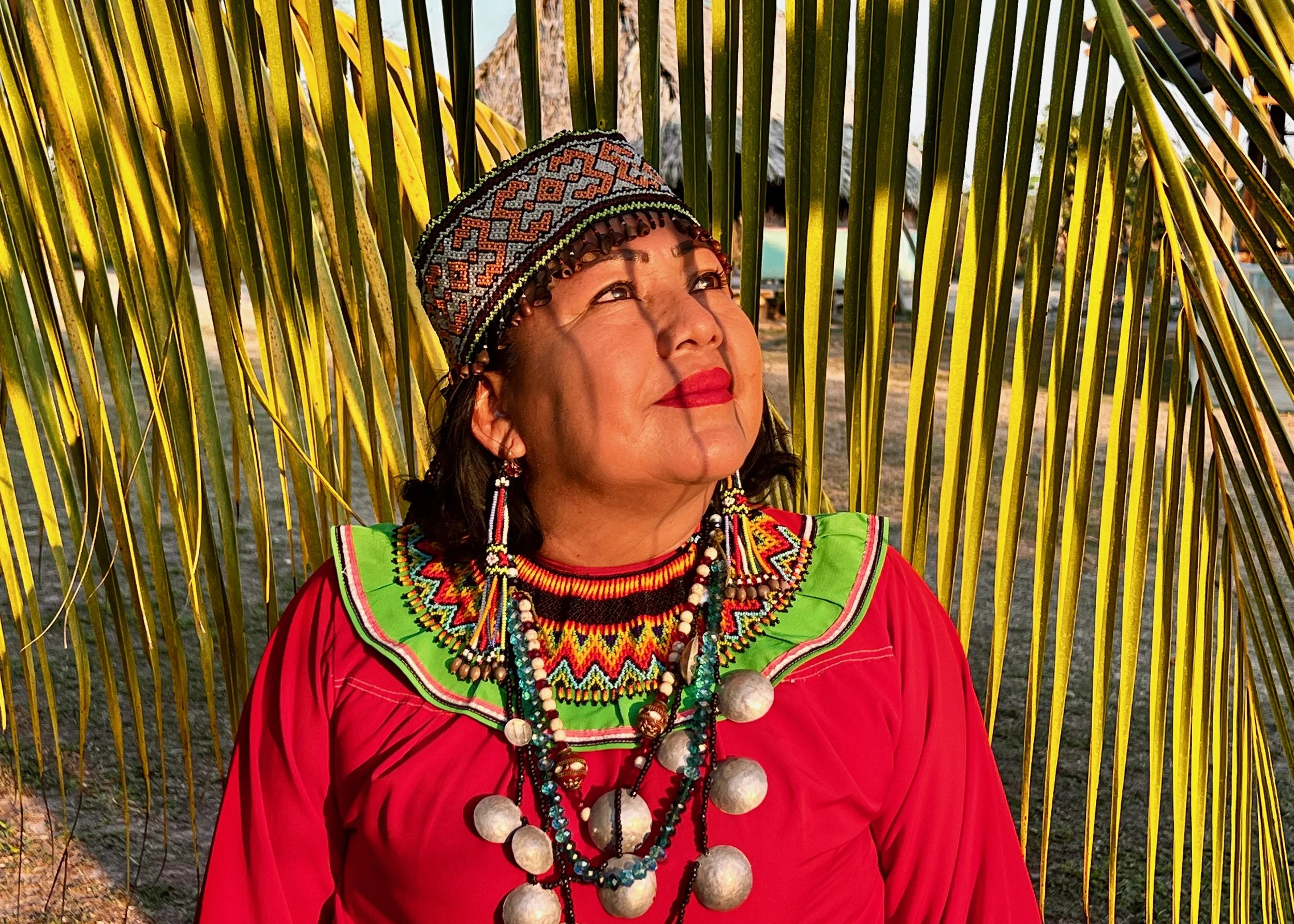 A woman wearing traditional indigenous jewelry and clothing, including a colorful beaded necklace and earrings, a patterned headscarf, and a red blouse, stands in front of tropical palm leaves on a beach at sunset.