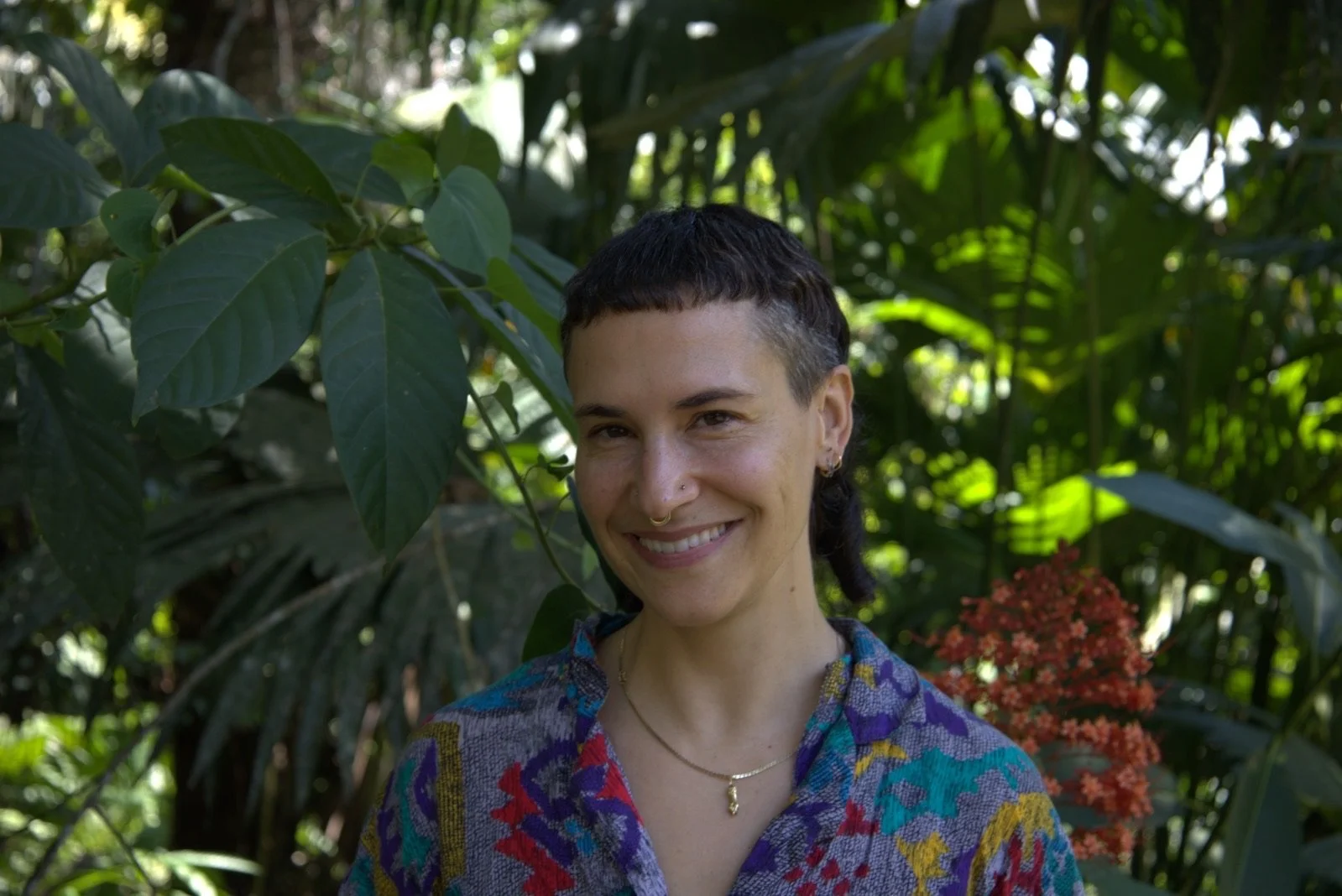 A person with short dark hair and a nose piercing, smiling, standing in lush green foliage, wearing a colorful patterned shirt and a gold necklace.