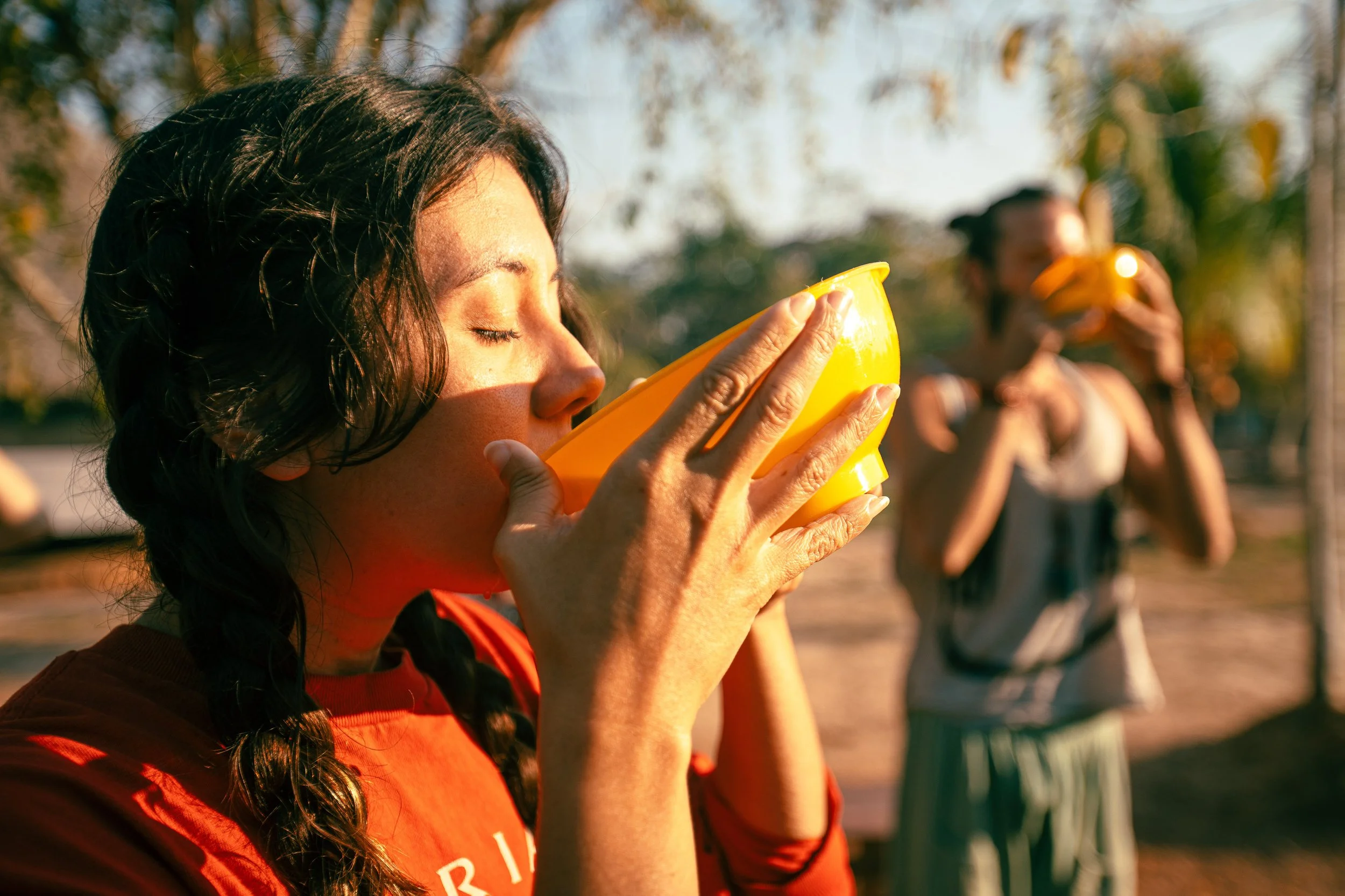 A woman in a red shirt drinks from a yellow bowl during a sunny outdoor setting, with another woman in the background also drinking from a yellow bowl.