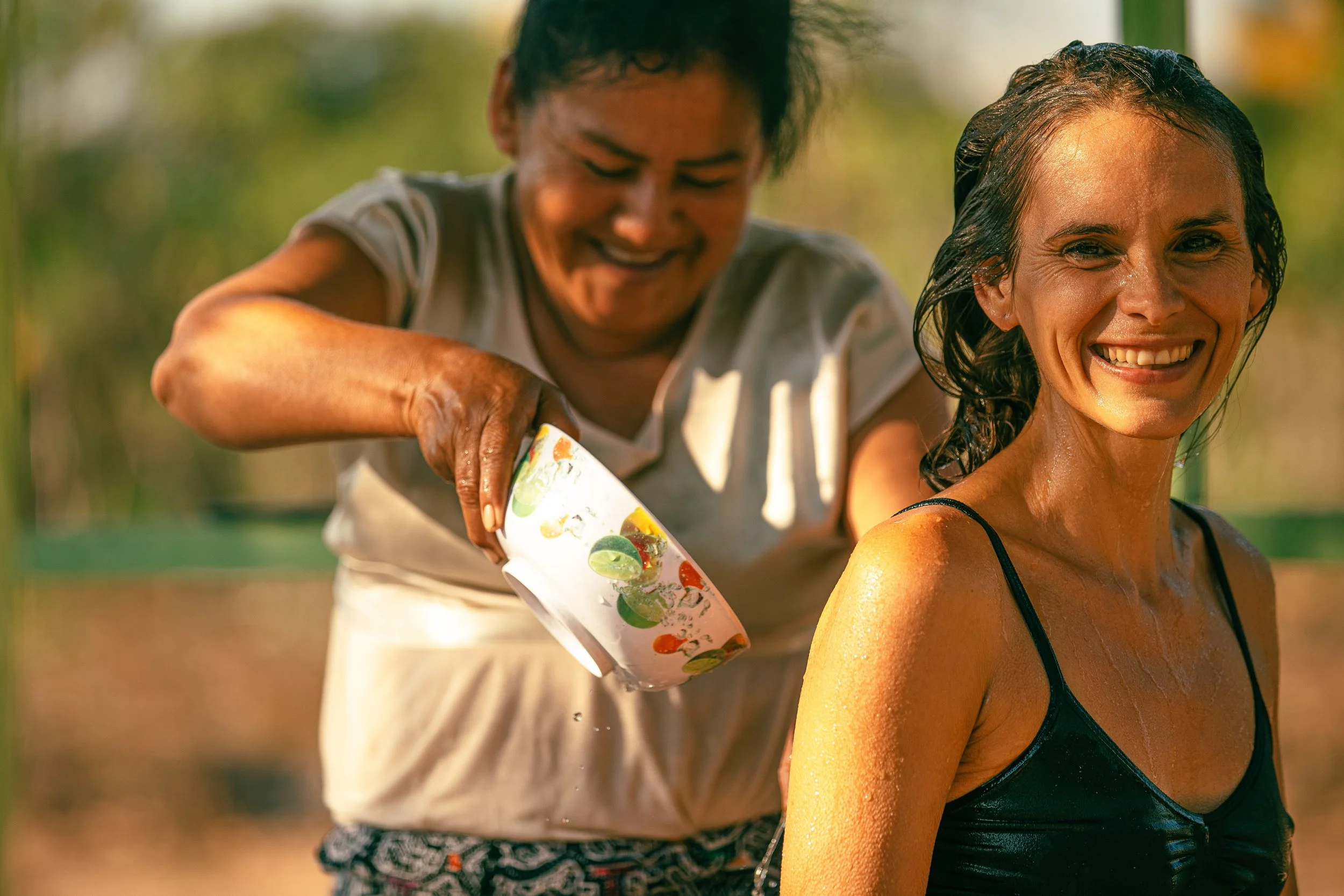 A woman in a black swimsuit smiling as she gets cold water poured over her from a colorful cup by another woman outdoors.