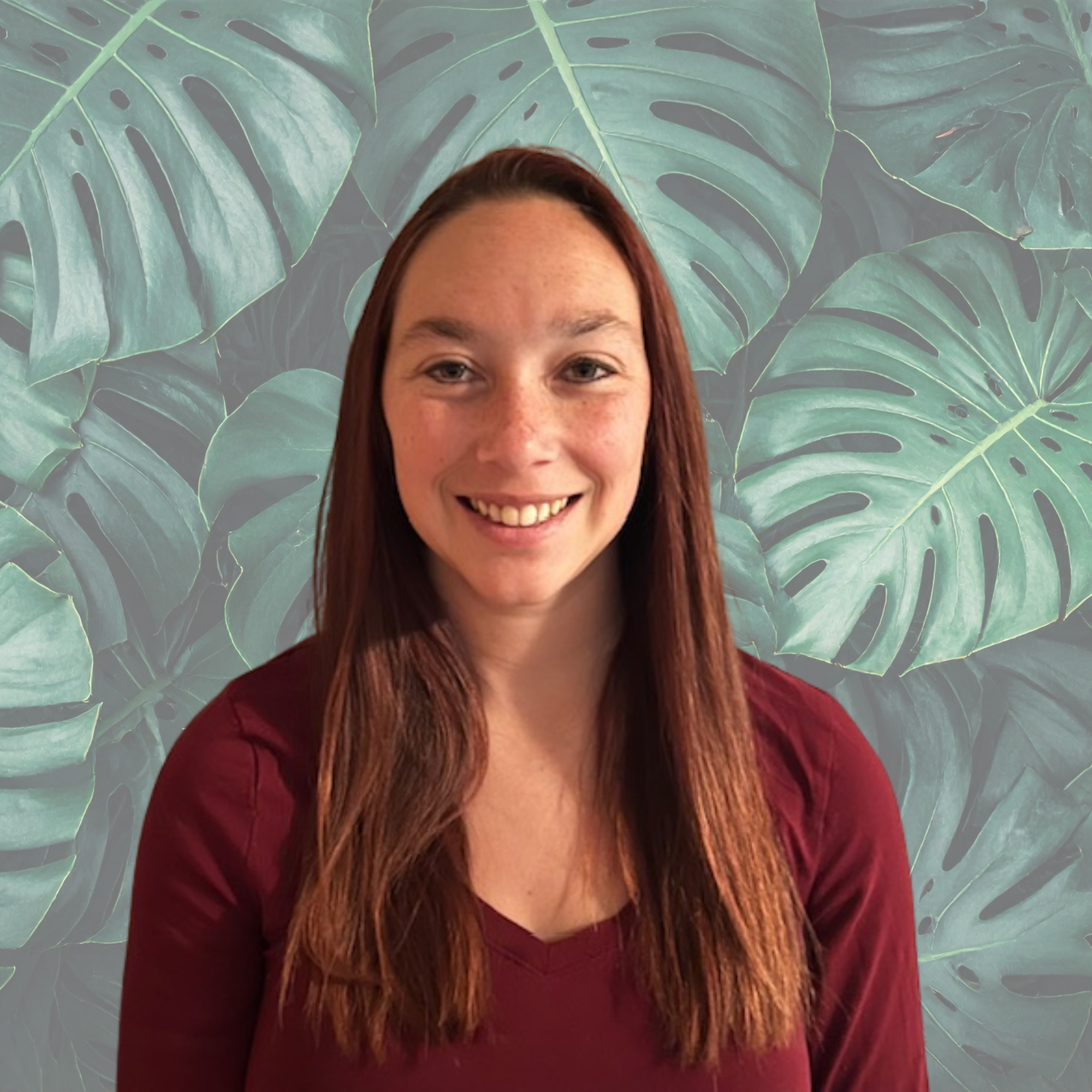 A young woman with long brown hair smiling at the camera, wearing a maroon top, standing against a plain light-colored wall.