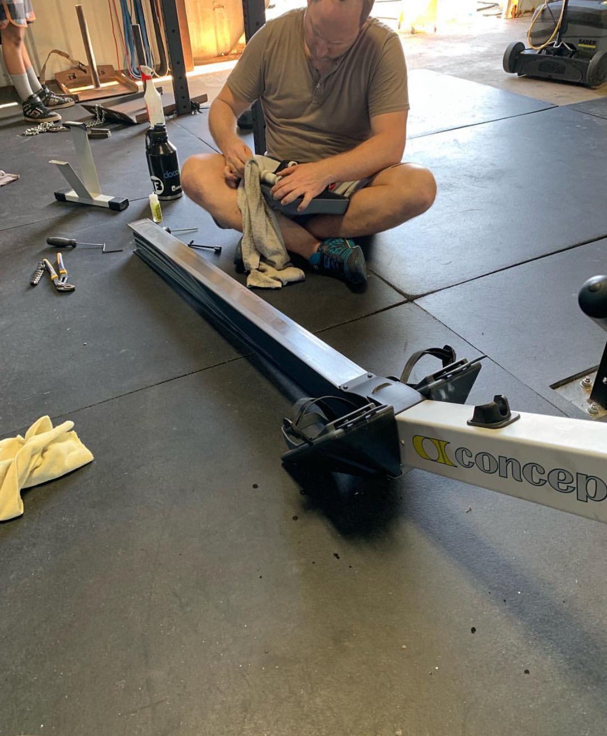A man sitting cross-legged on a garage floor working on a Concept2 indoor rowing machine. Various tools and a water bottle are around him.