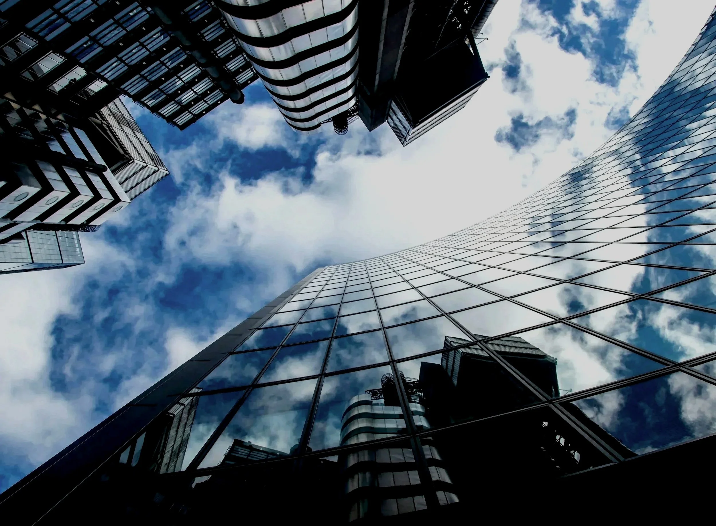 Low-angle view of modern glass skyscrapers with reflective windows, reaching towards a partly cloudy blue sky.