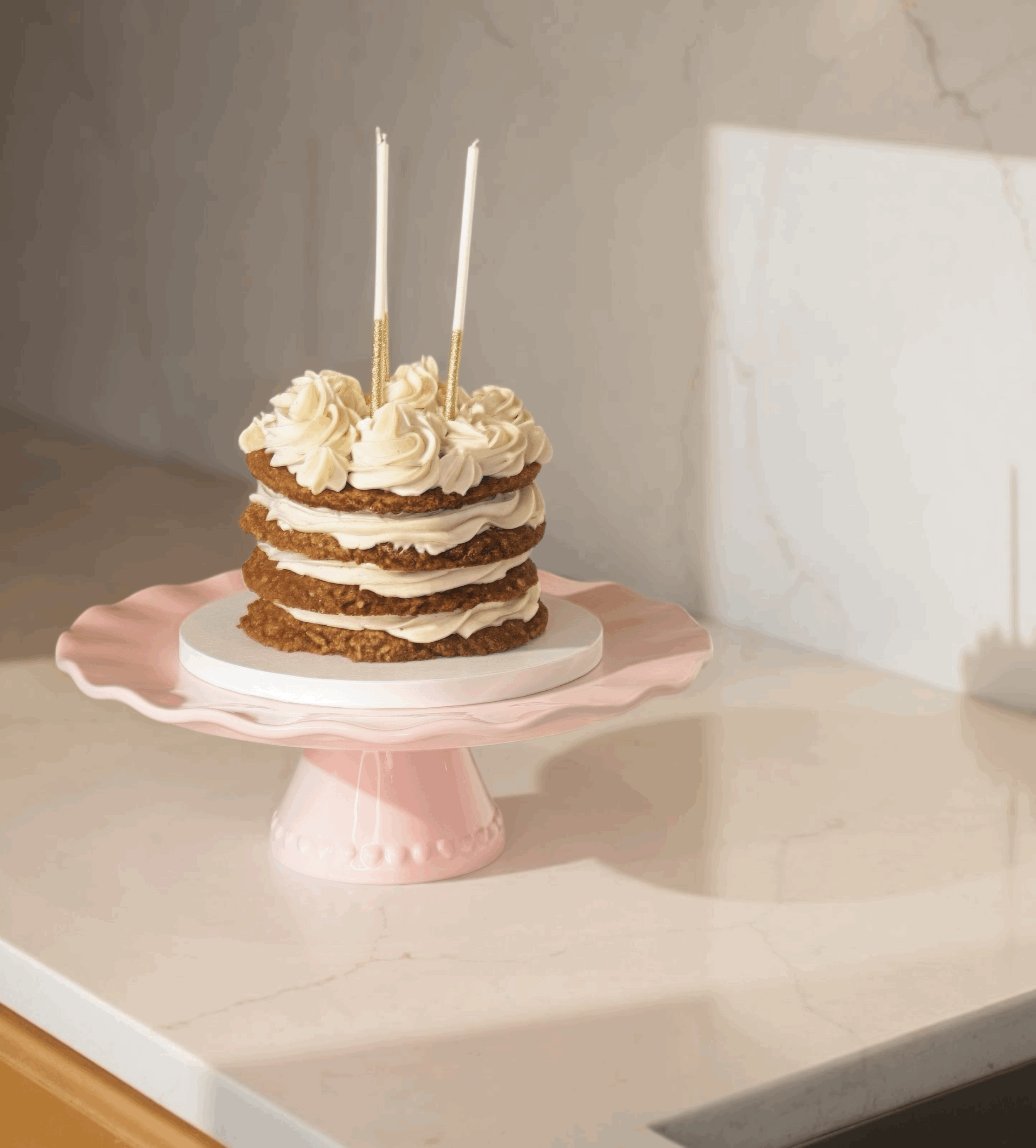 A layered cake with cookies and cream frosting on a pink pedestal cake stand, decorated with two white candles with gold accents, on a white marble countertop.