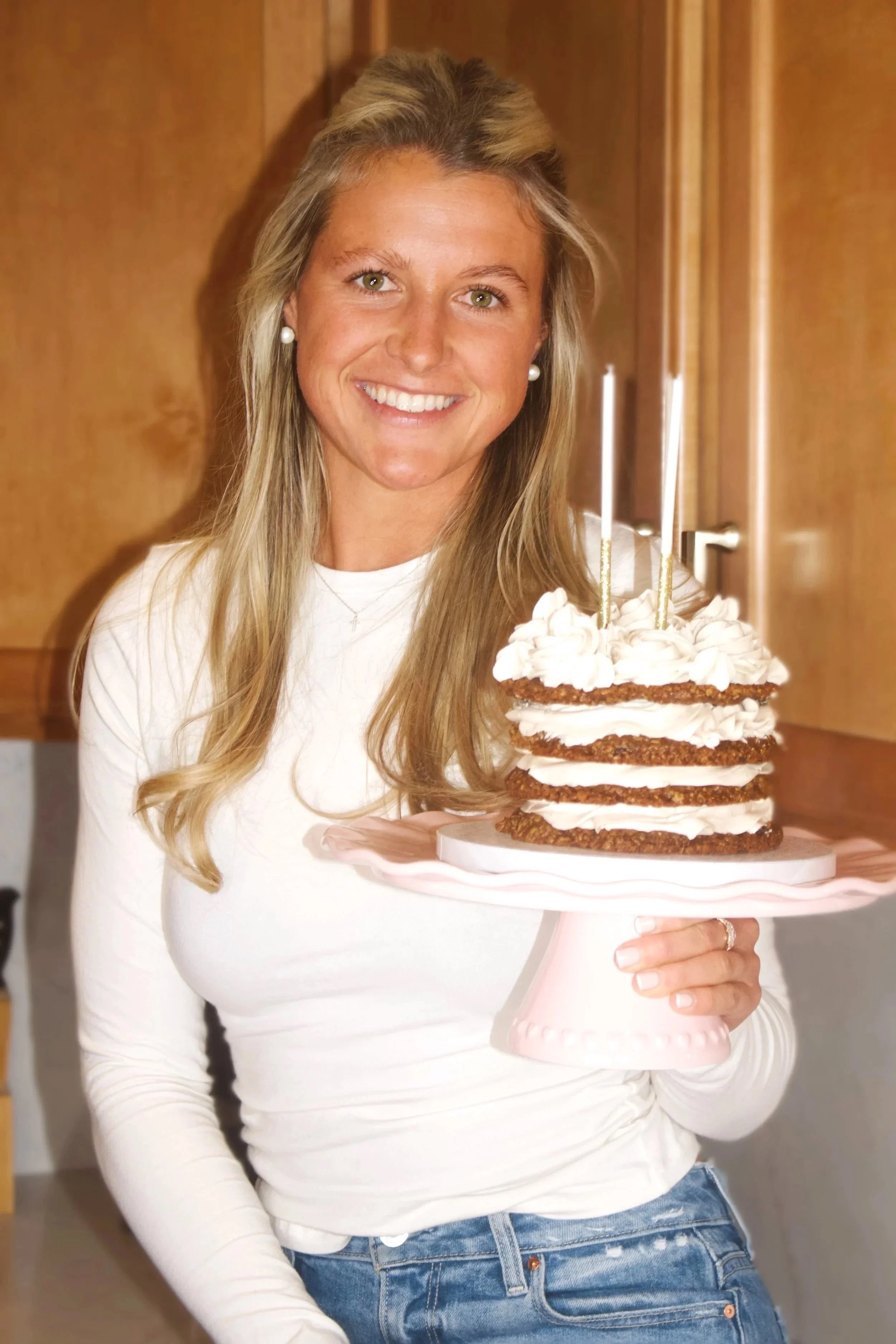 Smiling woman in a white long sleeve shirt and jeans holding a layered cake with white frosting on a pink cake stand in a kitchen.