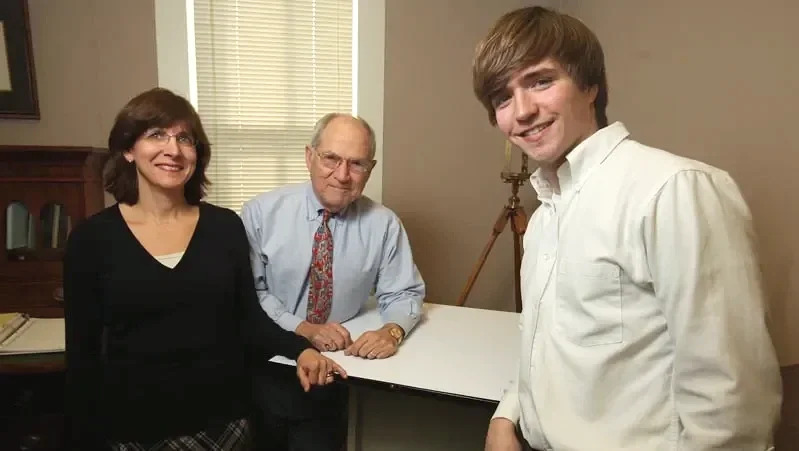 Three people, two women and one man, standing in a room with a piano and window blinds behind them.