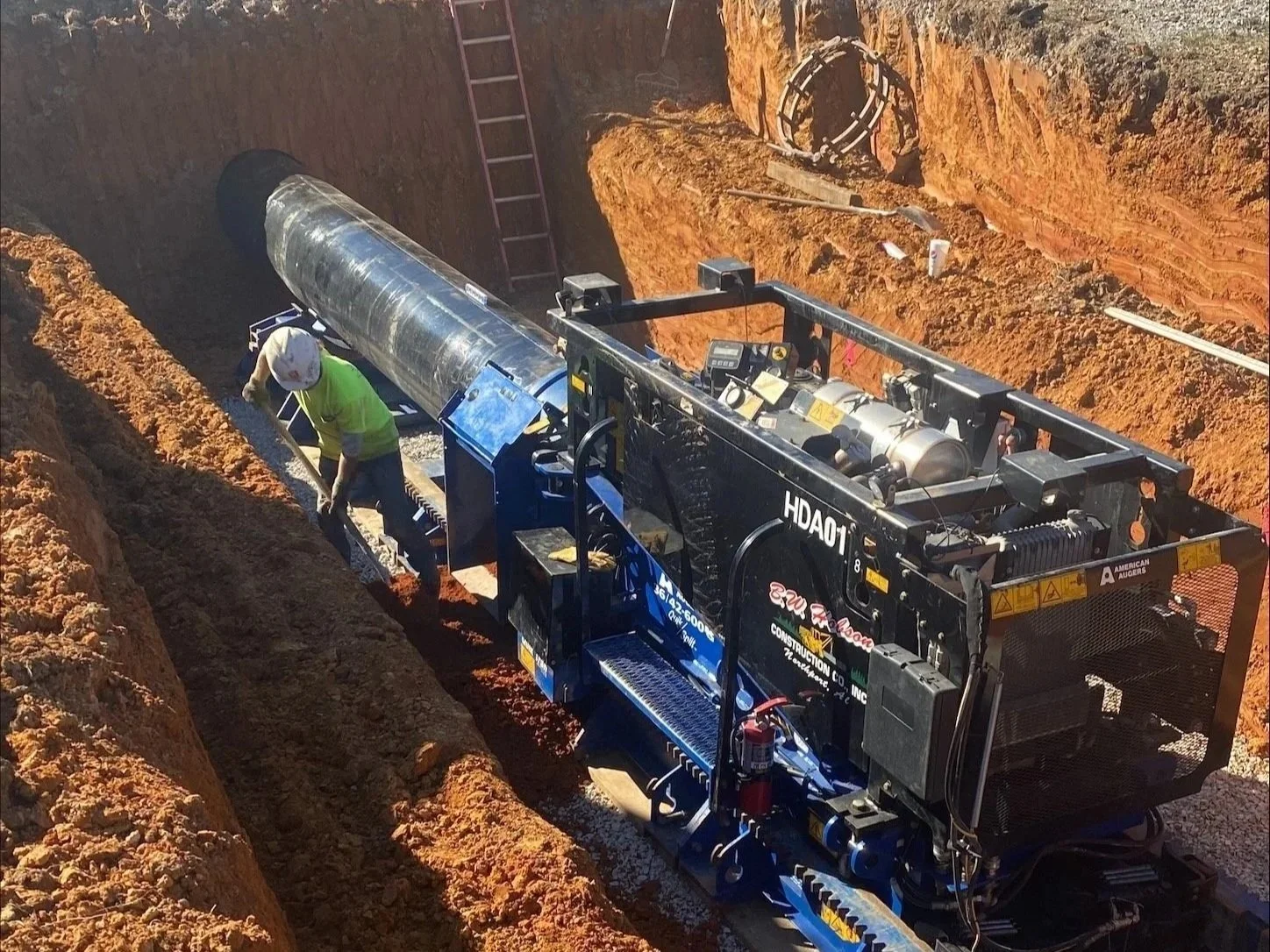 Construction worker installing a large metal pipe in a trench with a specialized pipe-laying machine.