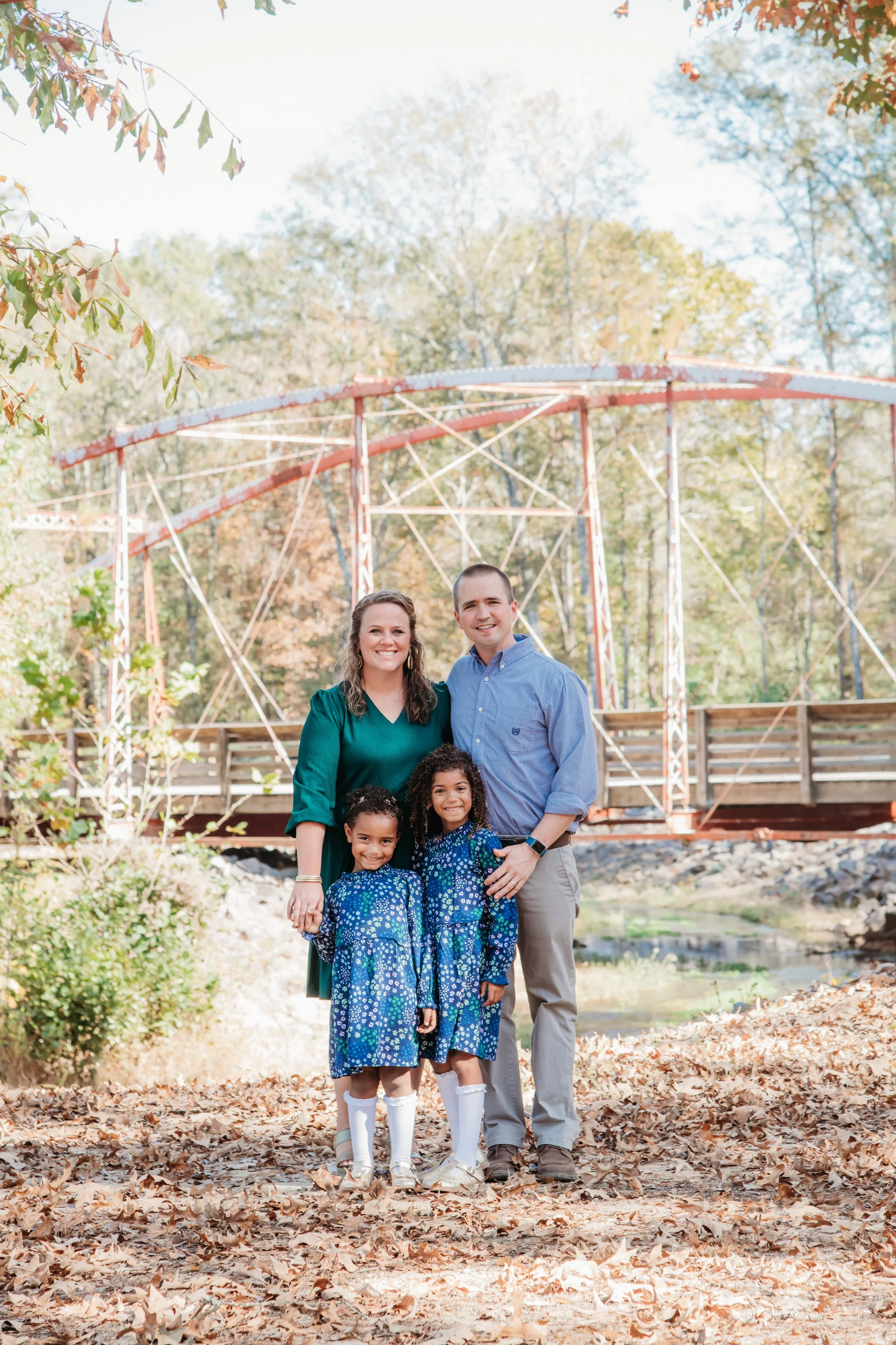 A family of four standing outdoors in a park with fallen leaves, posing in front of an old orange and white roller coaster. The family includes a mother, father, and two young daughters, all smiling.