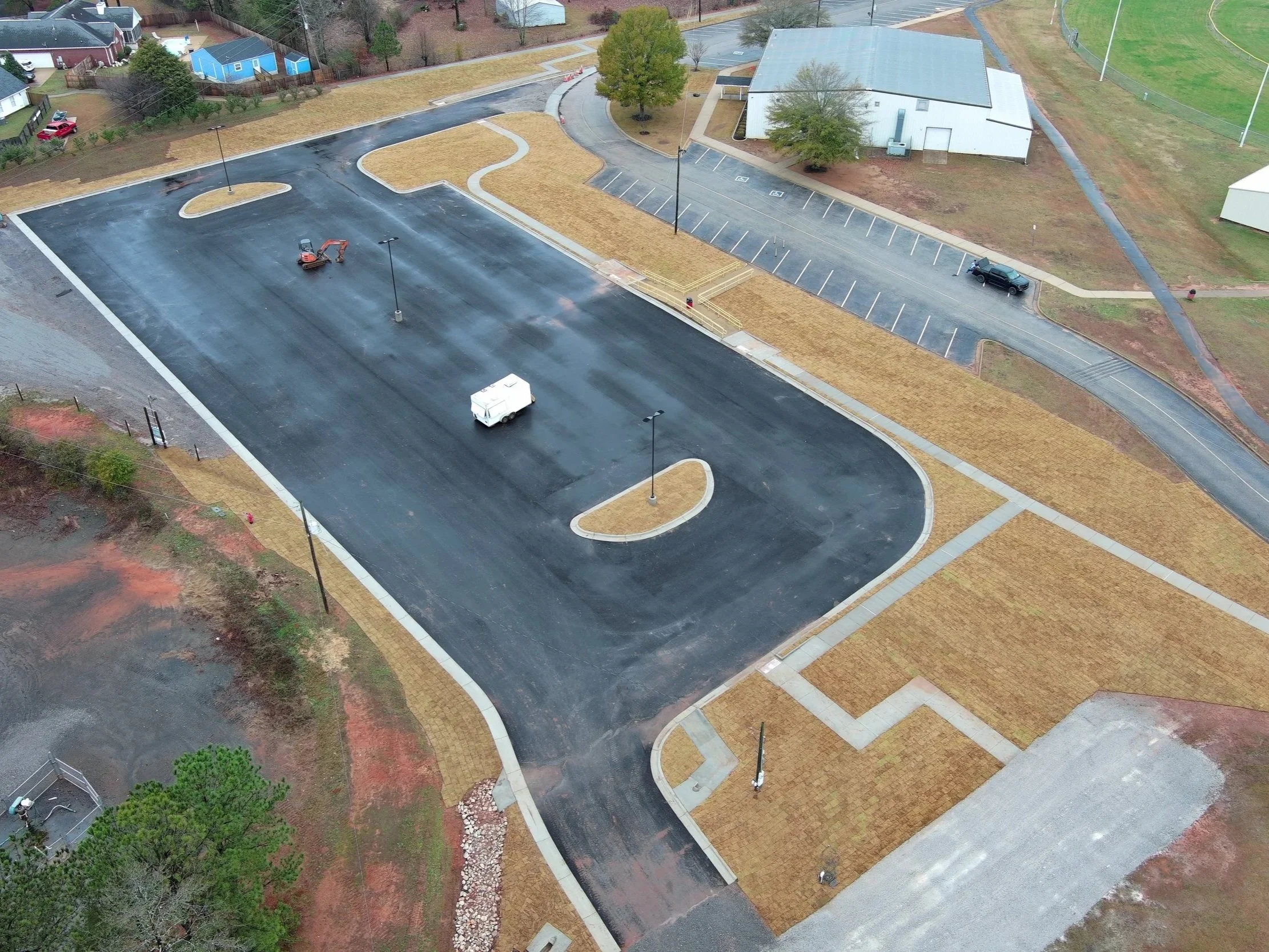Empty parking lot with a small construction vehicle and a white utility van. Surrounding area includes some trees, grass, and sidewalk paths. Nearby a building and parked car are visible.
