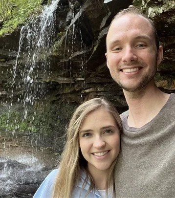 A smiling man and woman taking a selfie outdoors near a small waterfall with rocks.