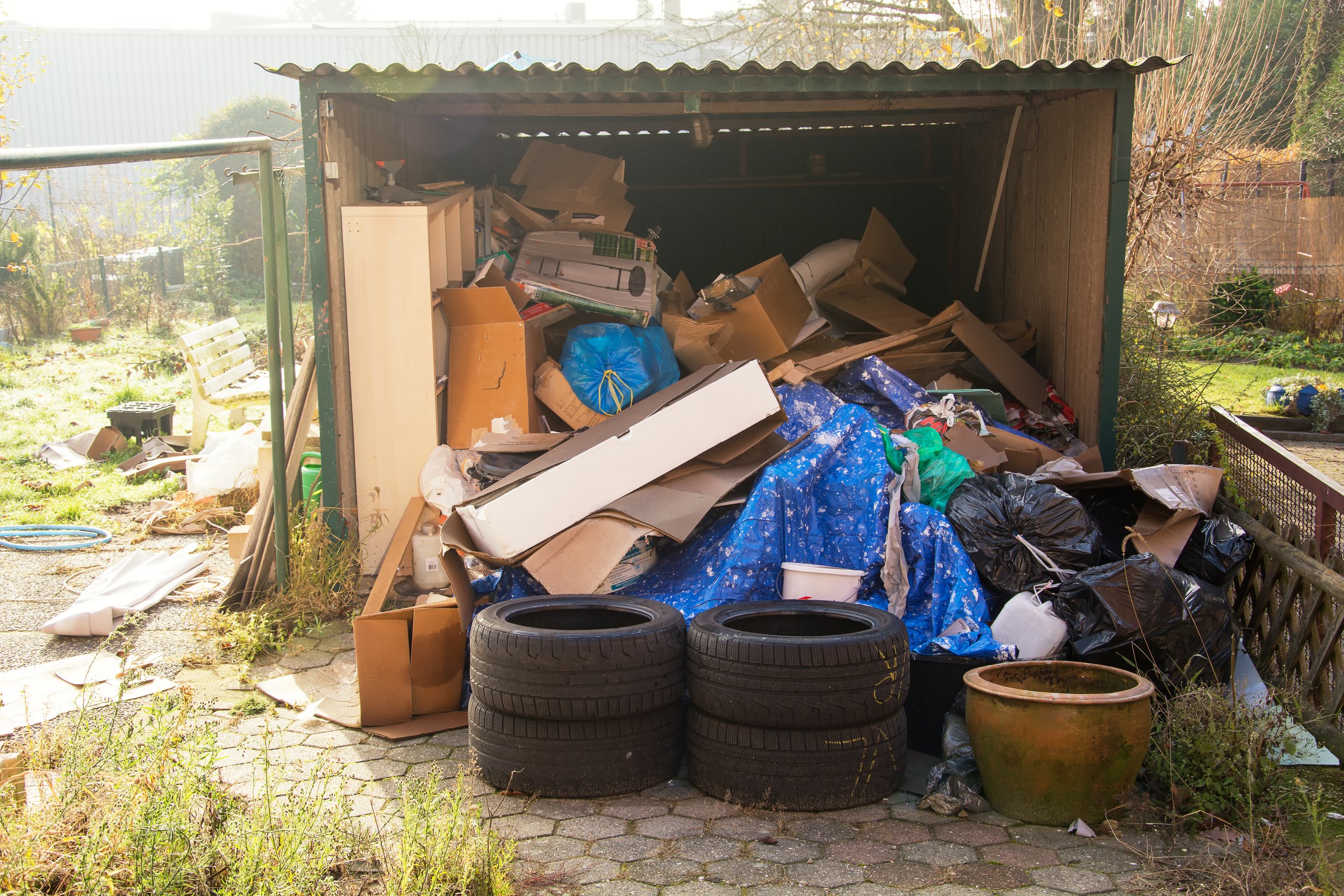 A cluttered outdoor storage shed filled with discarded cardboard, plastic, and other debris, with some tires and a large plant pot in front.