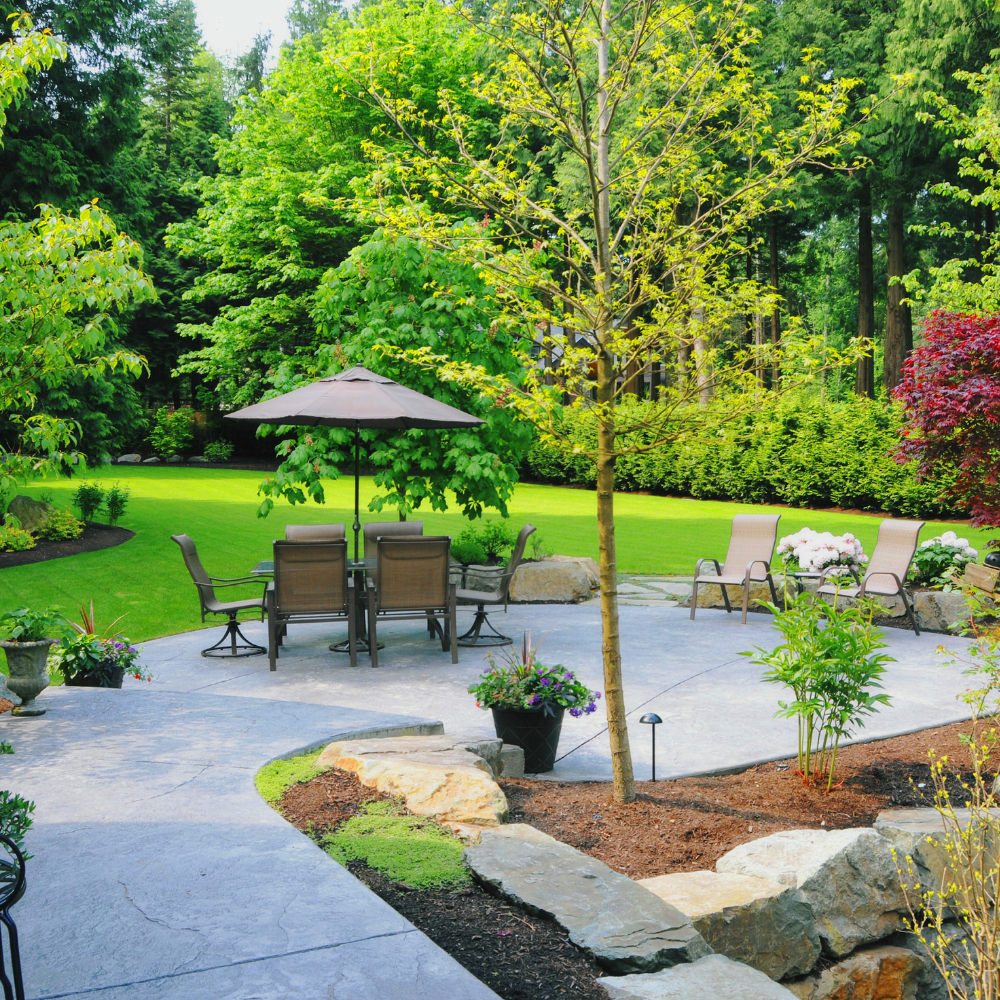 A backyard with a patio, a round outdoor dining table with six chairs and an umbrella, and two lounge chairs, with a lush green lawn and trees in the background.