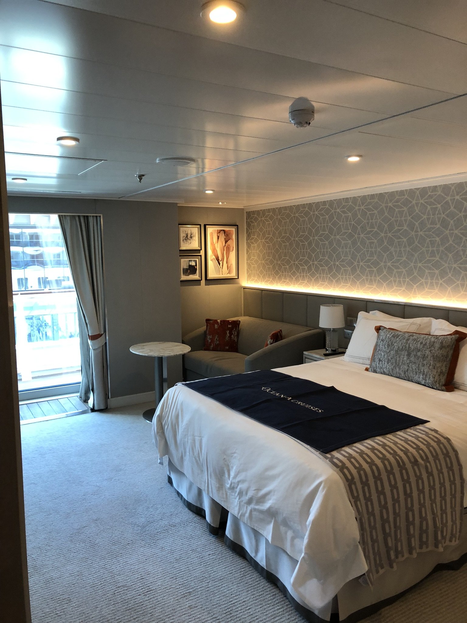 Interior of a cruise ship room with a bed, a sofa, a small table, a window with curtains, and framed artwork on the wall.
