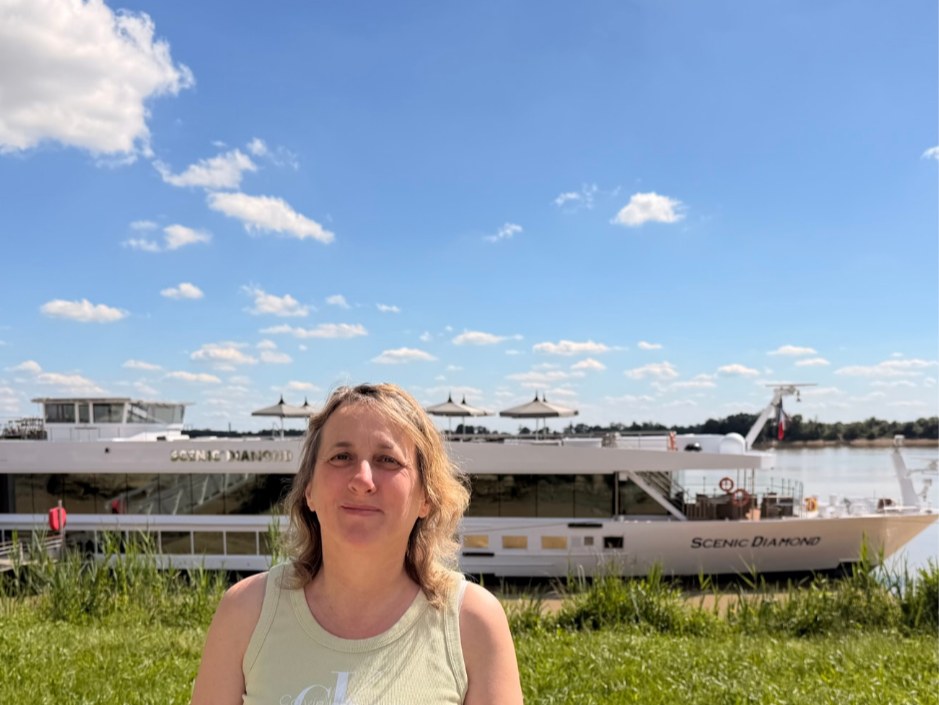 A woman standing outdoors near a river, with a large white boat named Scenic Diamond in the background under a blue sky with scattered clouds.