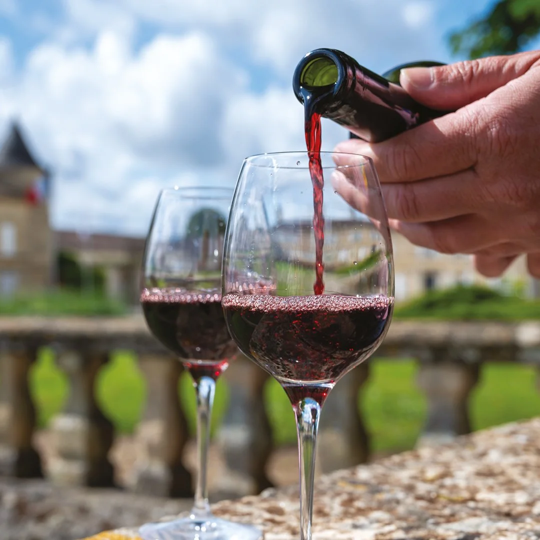 A hand pouring red wine into a wine glass on an outdoor table, with another wine glass filled with red wine in the background, against a partly cloudy sky and blurred buildings.