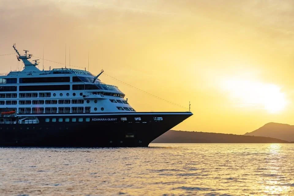 A large cruise ship named Azamara Quest sailing on calm water during sunset, with a mountainous landscape in the background.