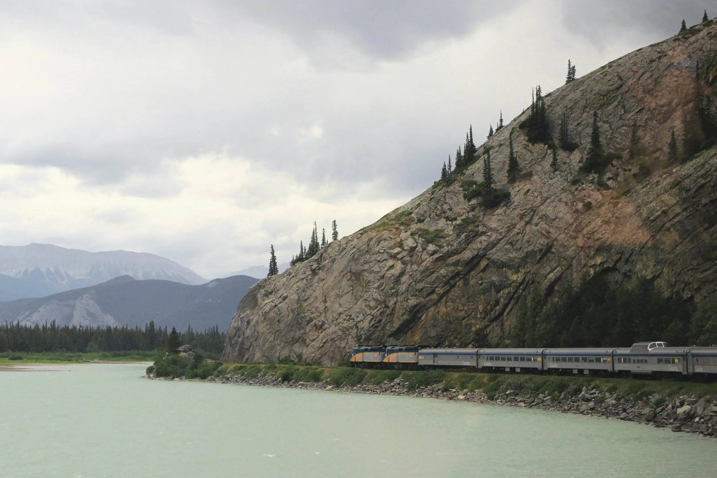 A train traveling along a railway track beside a green lake, with large rocky mountains and a cloudy sky in the background.