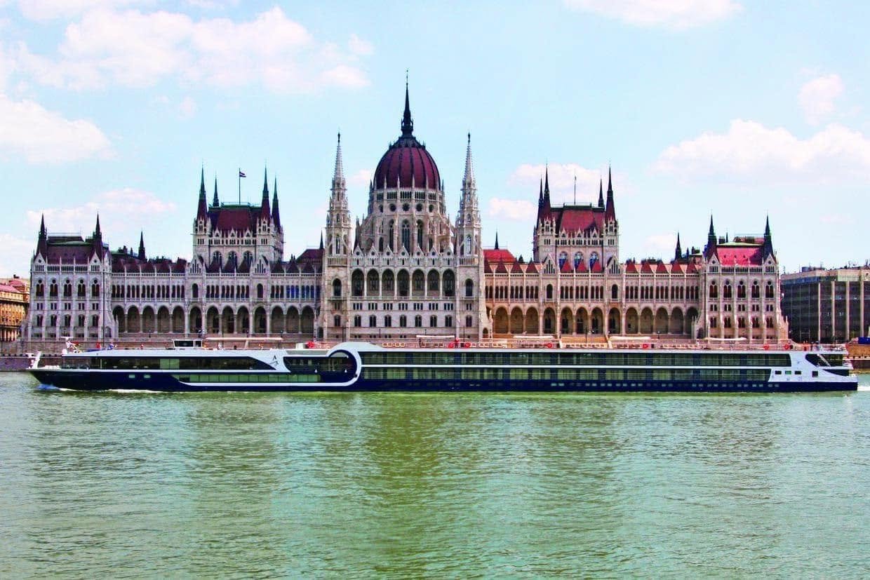 A large cruise ship sailing in a river, with the Parliament Building of Budapest, Hungary, in the background.