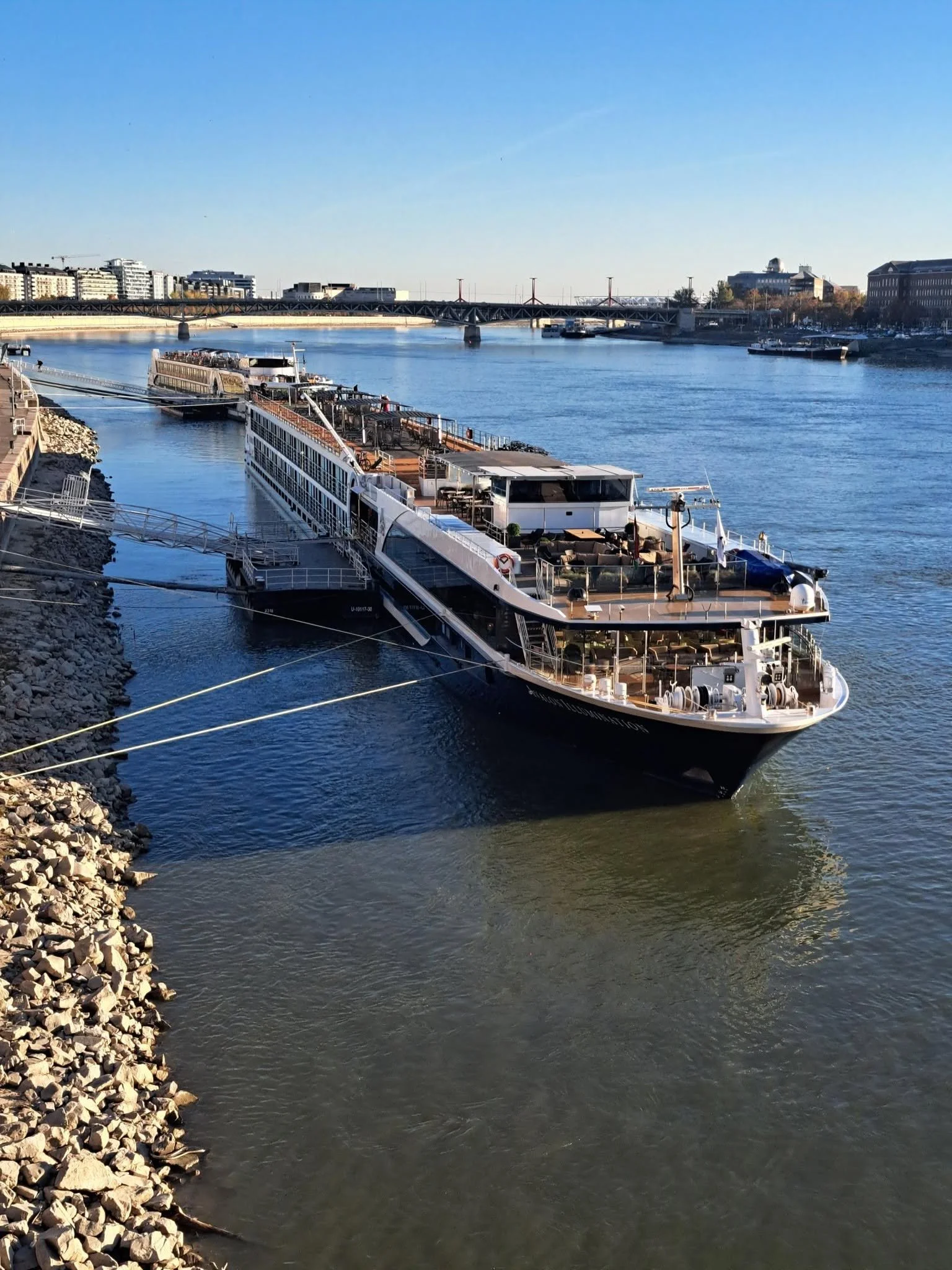 A river with a white cruise ship docked along the rocky shore, a bridge crossing the river, and buildings in the background on a clear day.