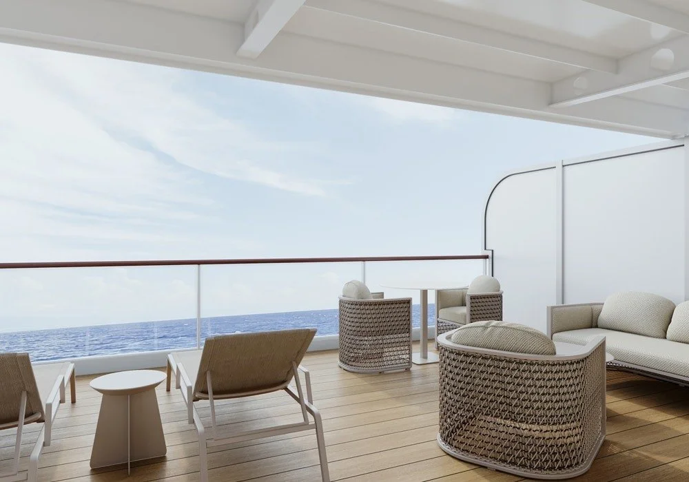 A spacious balcony with outdoor furniture overlooking the ocean, including two lounge chairs, a sitting area with a sofa and a round table, and two woven chairs, all under a partly covered roof.